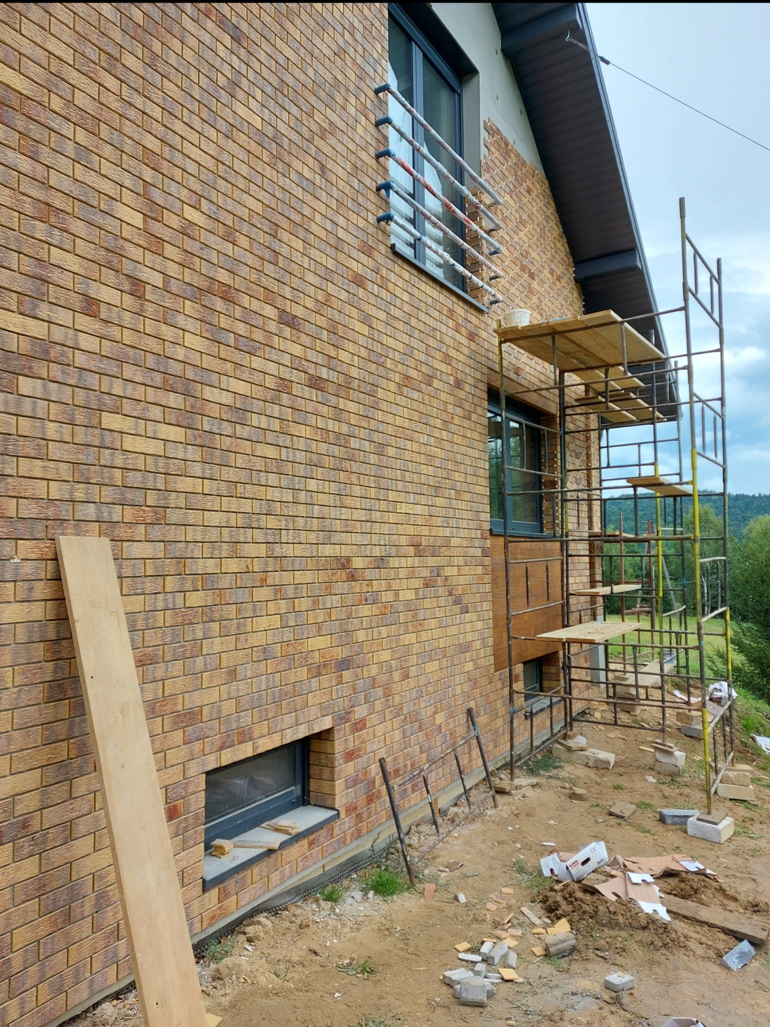 Dom w trakcie wykańczania elewacji z cegły klinkierowej, widoczne rusztowanie, okno z kratami i deska oparta o ścianę.