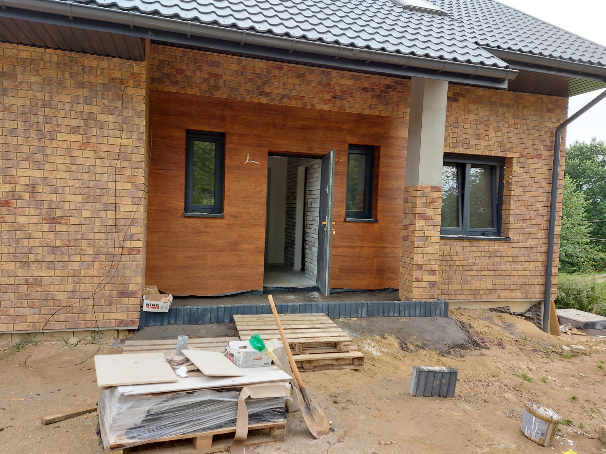 Nowo wybudowany dom z elewacją z cegły klinkierowej i drewnopodobną, czarnymi oknami, otwartymi drzwiami wejściowymi, widocznymi materiałami budowlanymi i narzędziami na pierwszym planie.