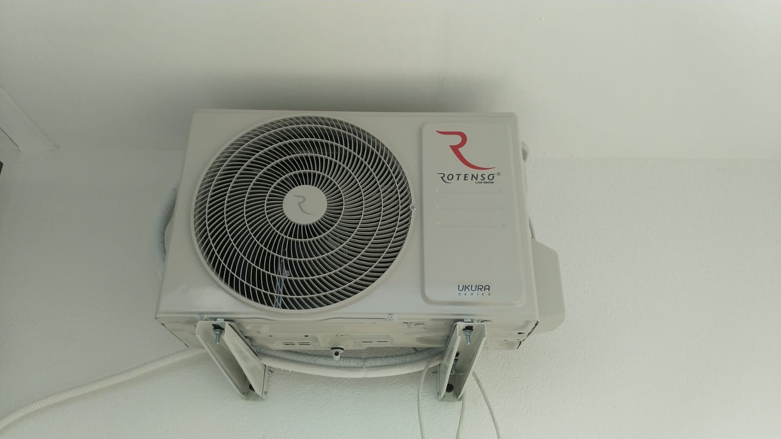 Biała jednostka zewnętrzna klimatyzacji Rotenso Ukura Series zamontowana na białej ścianie, widoczne elementy montażowe i przewody.