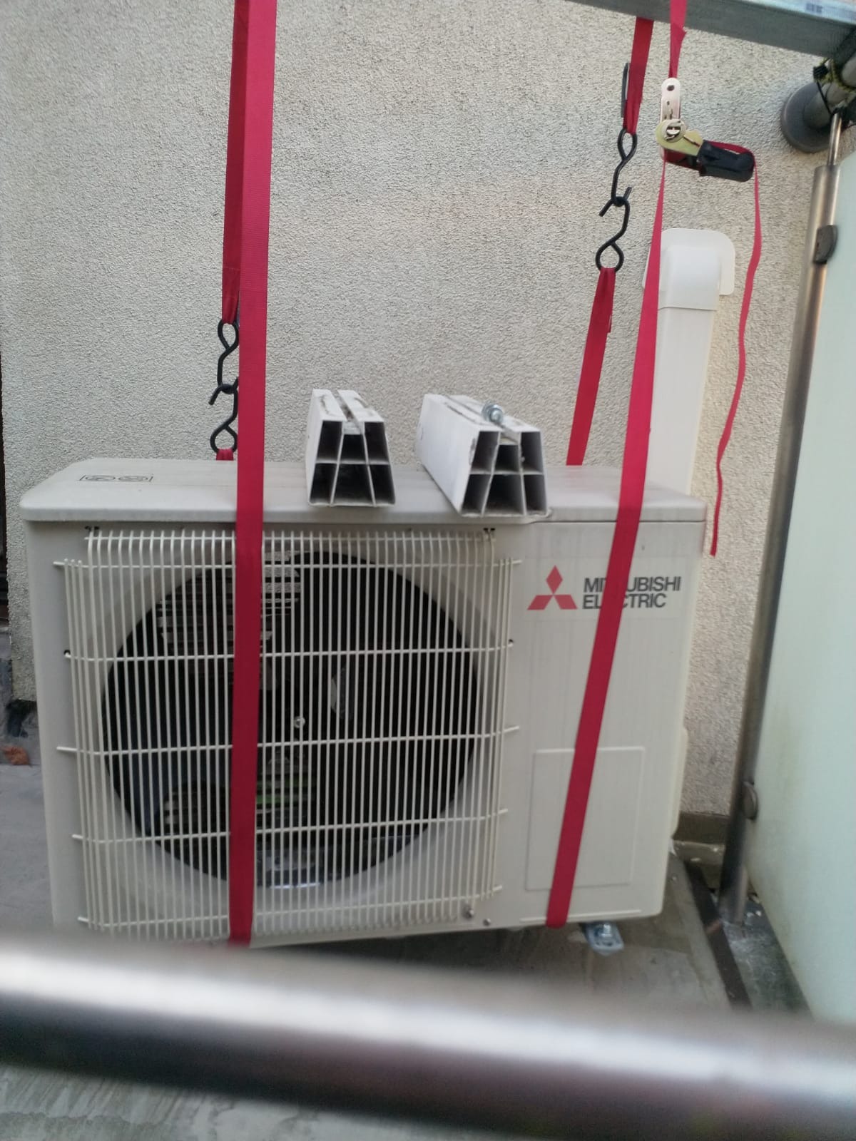 Zewnętrzna jednostka klimatyzacji Mitsubishi Electric zawieszona na czerwonych pasach transportowych, z dwoma aluminiowymi profilami na górze, gotowa do montażu.