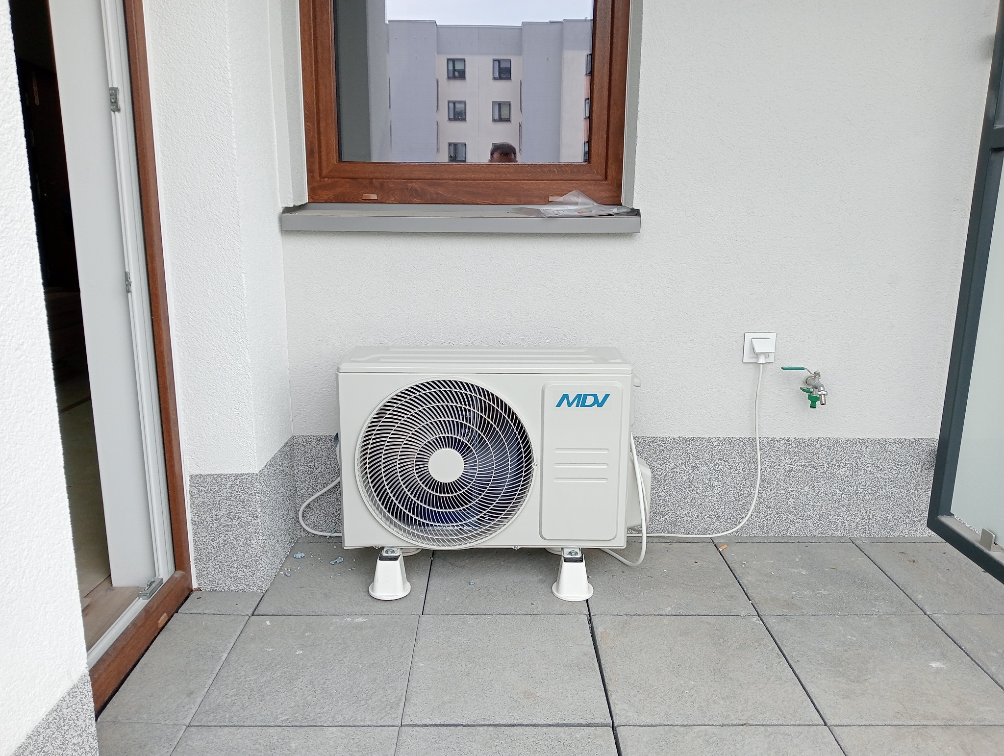 Biała jednostka zewnętrzna klimatyzacji MDV zamontowana na balkonie z widokiem na budynek mieszkalny odbijający się w oknie.