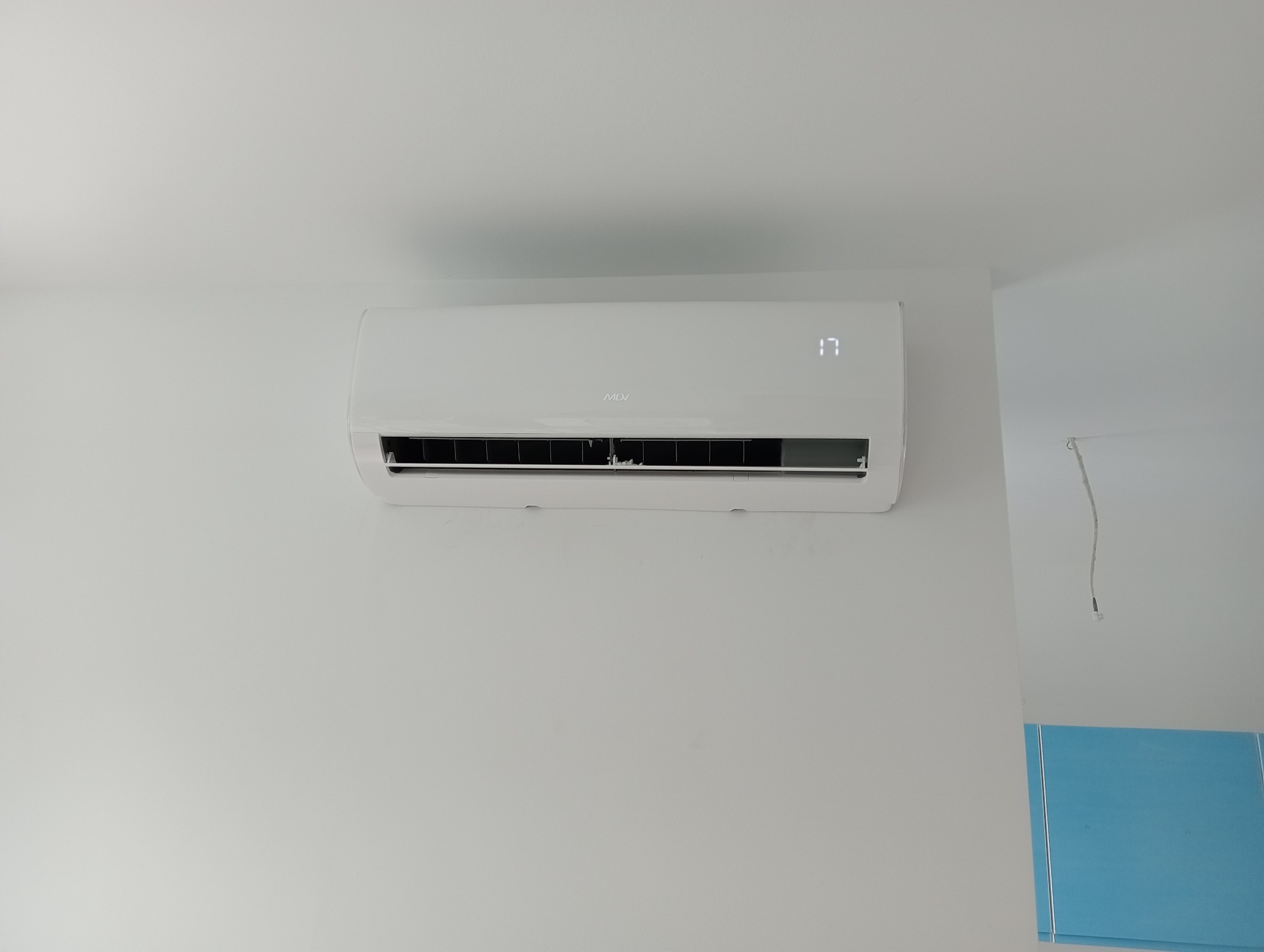 Biały klimatyzator MDV zamontowany na białej ścianie, wyświetlający temperaturę 17 stopni Celsjusza, z widocznym fragmentem niebieskiego zabezpieczenia na ścianie z prawej strony.
