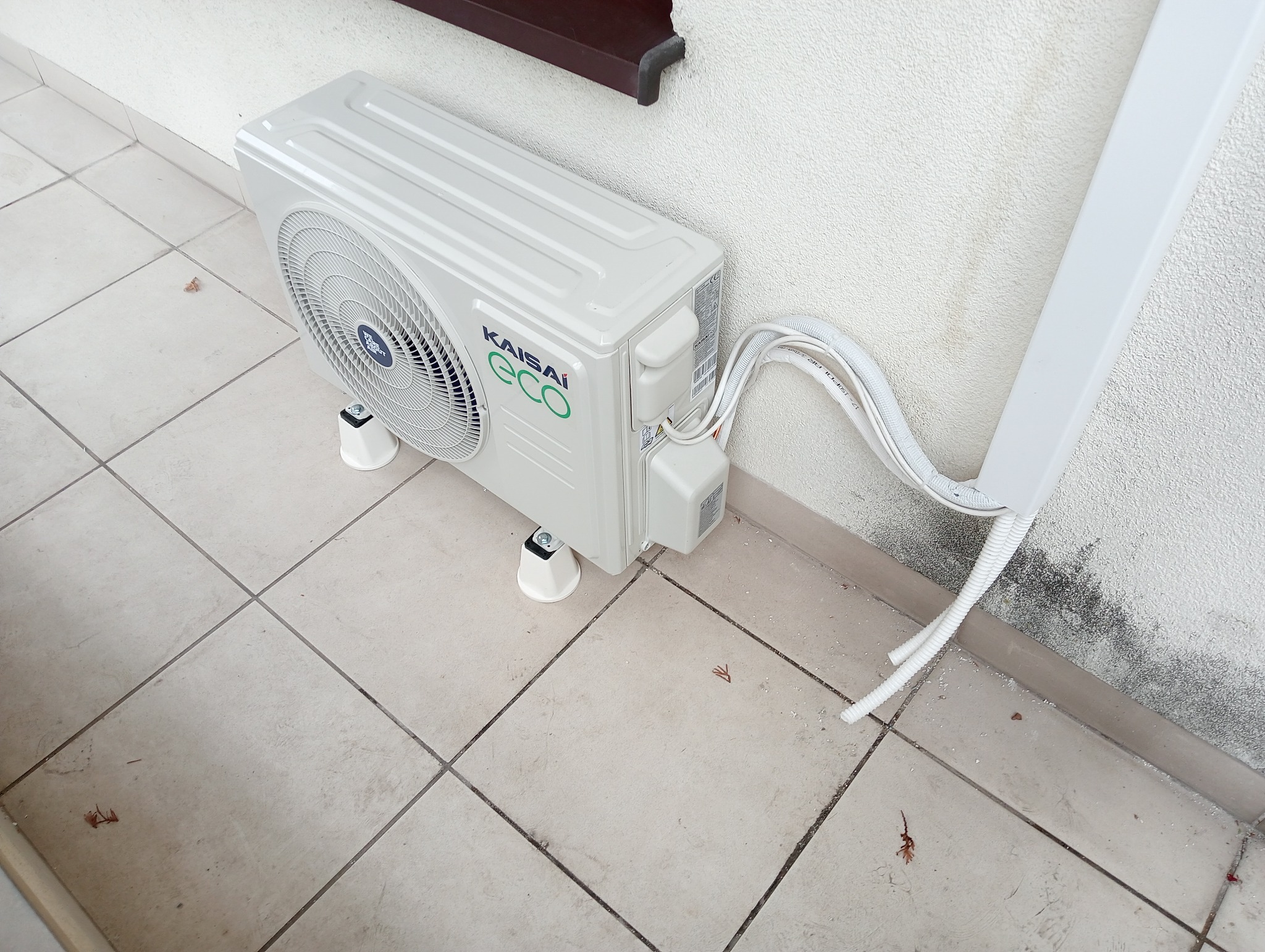 Zewnętrzna jednostka klimatyzacji Kaisai Eco zamontowana na białych wspornikach na płytkach, z widocznymi przewodami odprowadzającymi do ściany budynku.