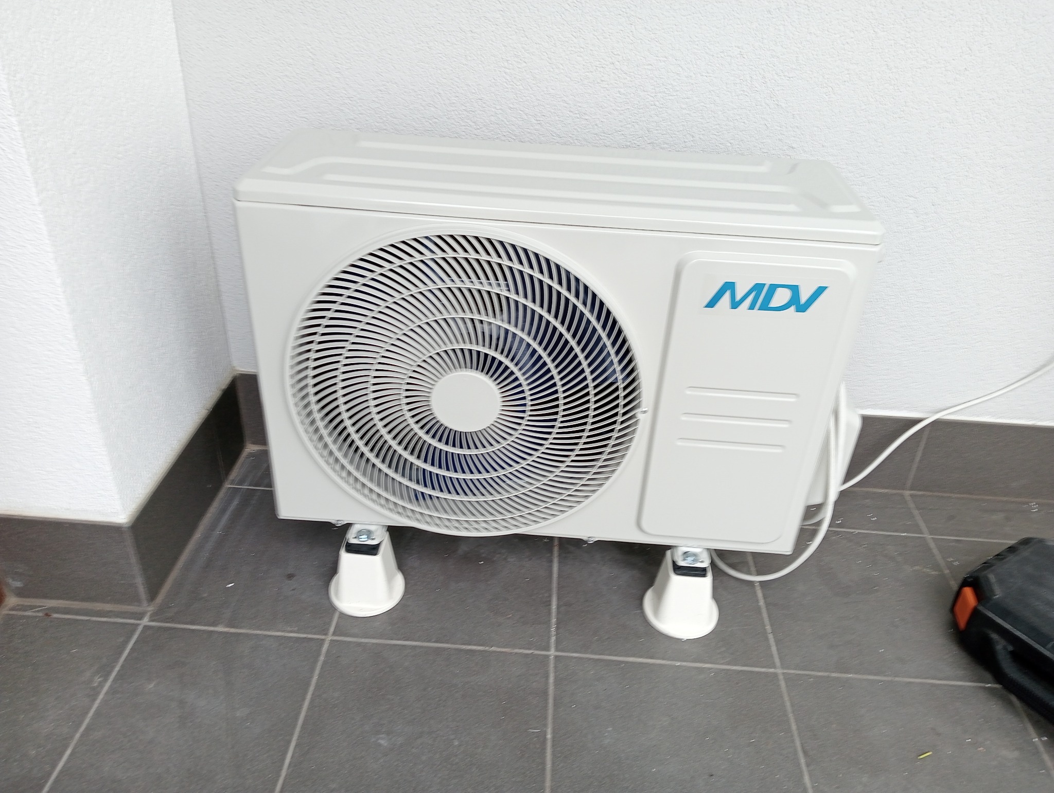 Biała zewnętrzna jednostka klimatyzacji MDV ustawiona na szarych płytkach pod ścianą, widoczny kabel zasilający i fragment czarnej skrzynki narzędziowej.