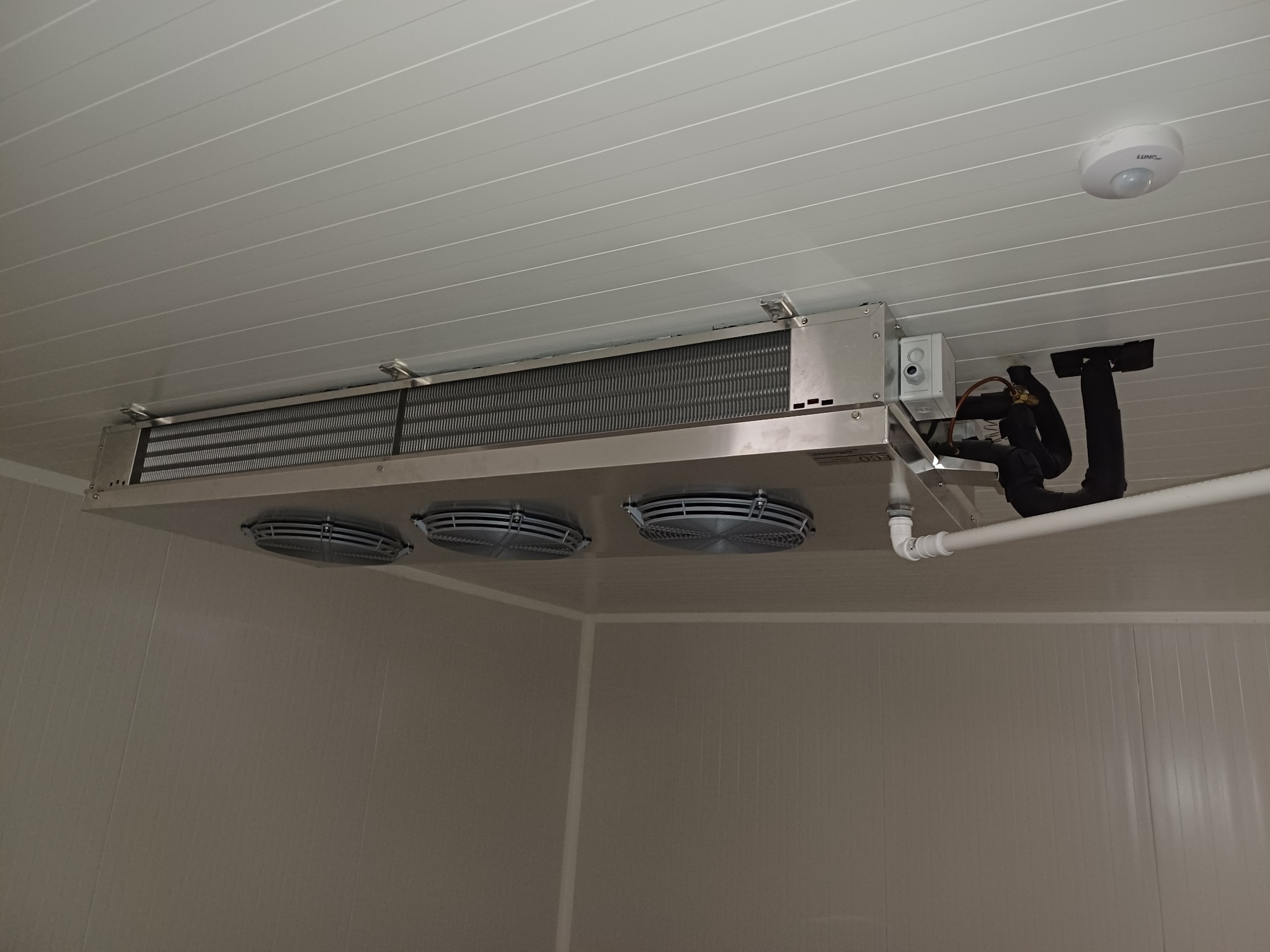Sufitowa chłodnica przemysłowa z trzema wentylatorami zamontowana w chłodni, widoczne rury i instalacja elektryczna.