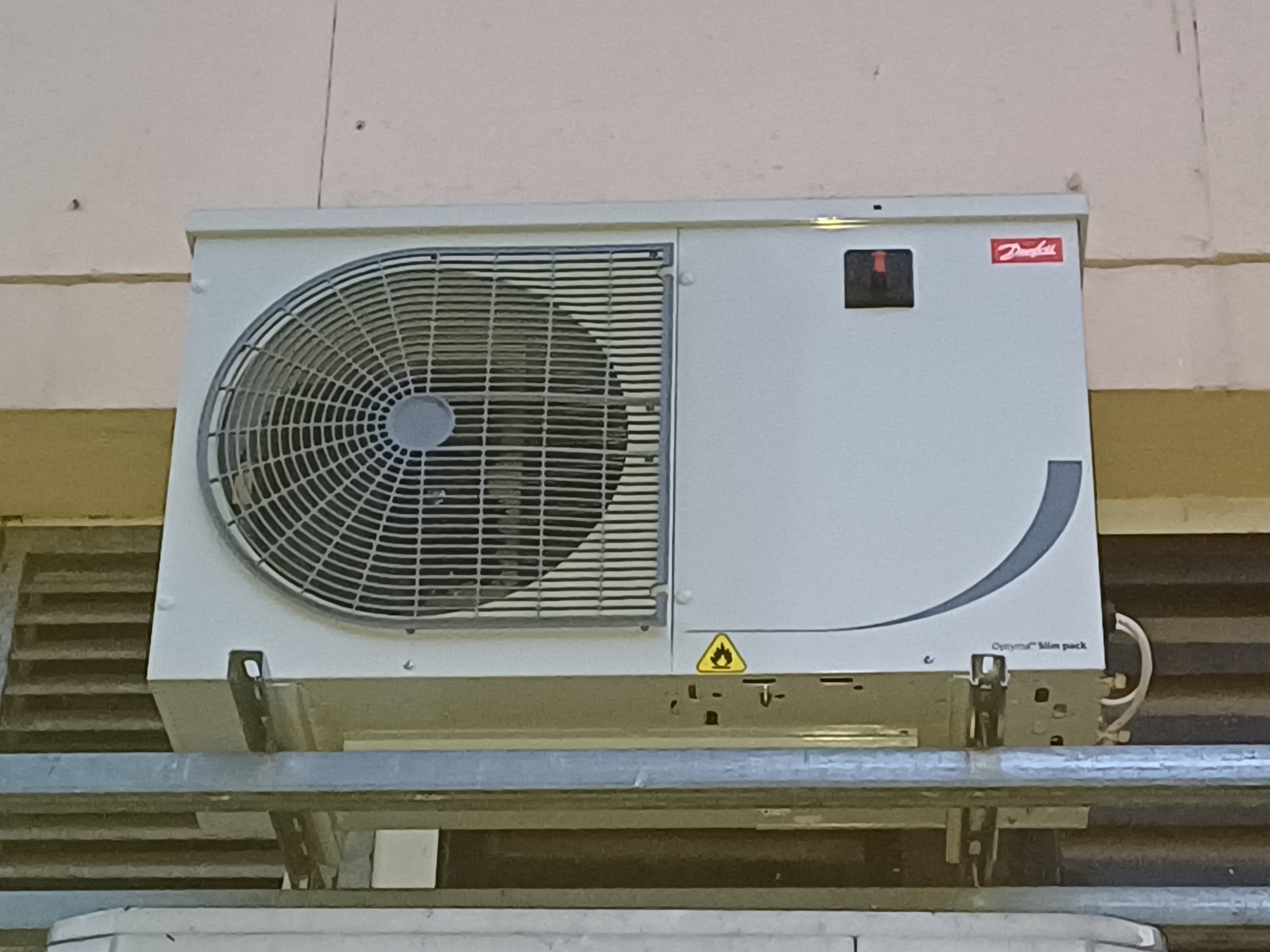 Biała jednostka zewnętrzna klimatyzacji Danfoss zamontowana na metalowej konstrukcji wsporczej na tle ściany.