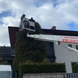 Anatolii Kononchuk Twój Ogrodnik - Dwaj pracownicy w podnośniku koszowym przycinają wysokie drzewo iglaste obok domu, widoczny biały bus i ogrodzenie z numerem posesji.