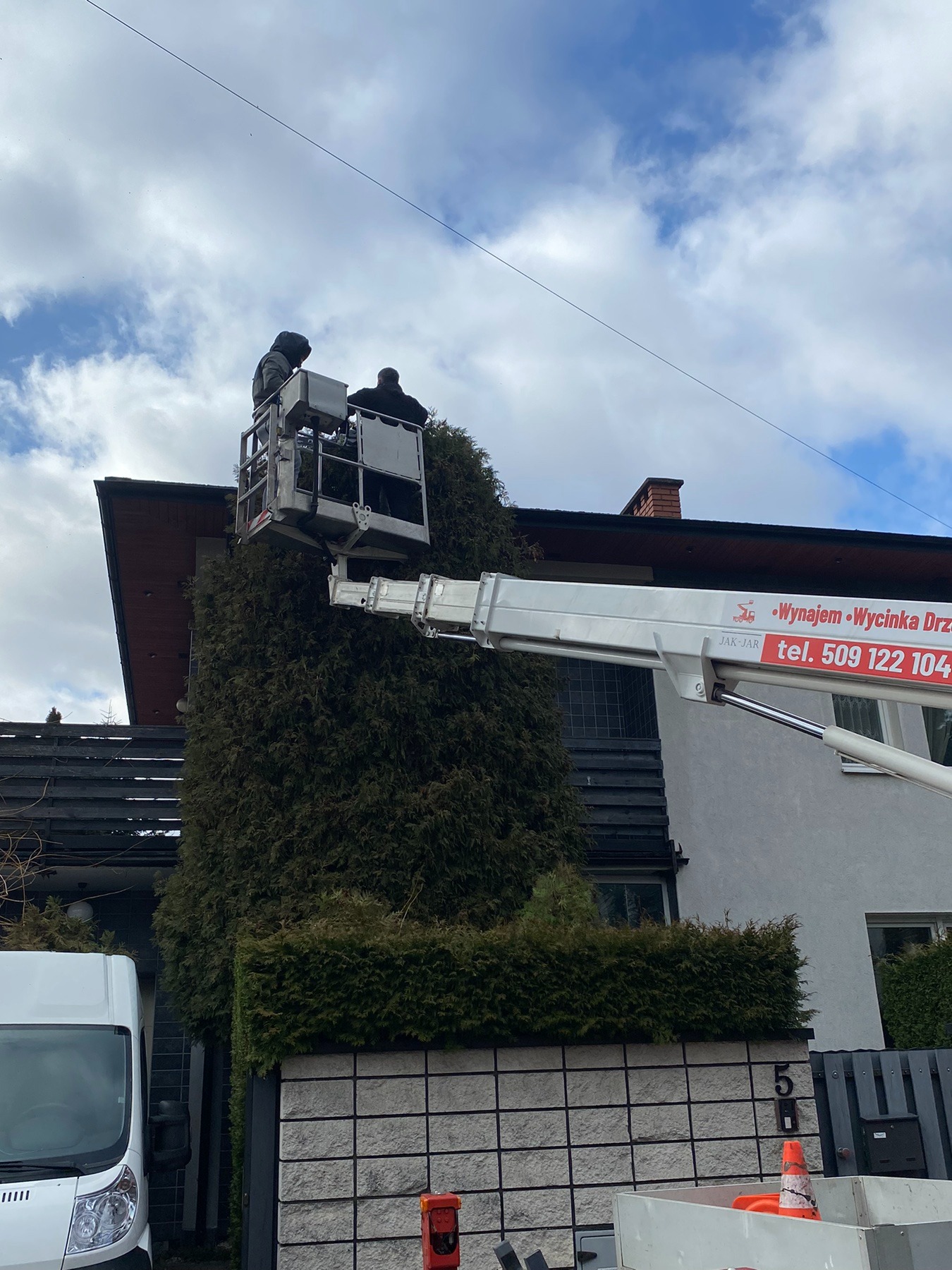 Dwaj pracownicy w podnośniku koszowym przycinają wysokie drzewo iglaste obok domu, widoczny biały bus i ogrodzenie z numerem posesji.