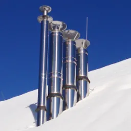 Cztery błyszczące, metalowe kominy wystające z zasypanego śniegiem dachu na tle intensywnego, błękitnego nieba.