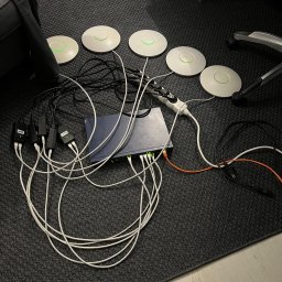 Hubert Zaczek - Rozłożone na ciemnoszarym dywanie: router, cztery białe punkty dostępowe z zielonymi diodami, zasilacze PoE i plątanina kabli sieciowych.