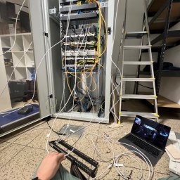 Hubert Zaczek - Otwarta szafa serwerowa z widocznymi patch panelami i switchami, niepodłączone kable, drabina aluminiowa, laptop z włączonym filmem i ręka z zegarkiem na pierwszym planie.