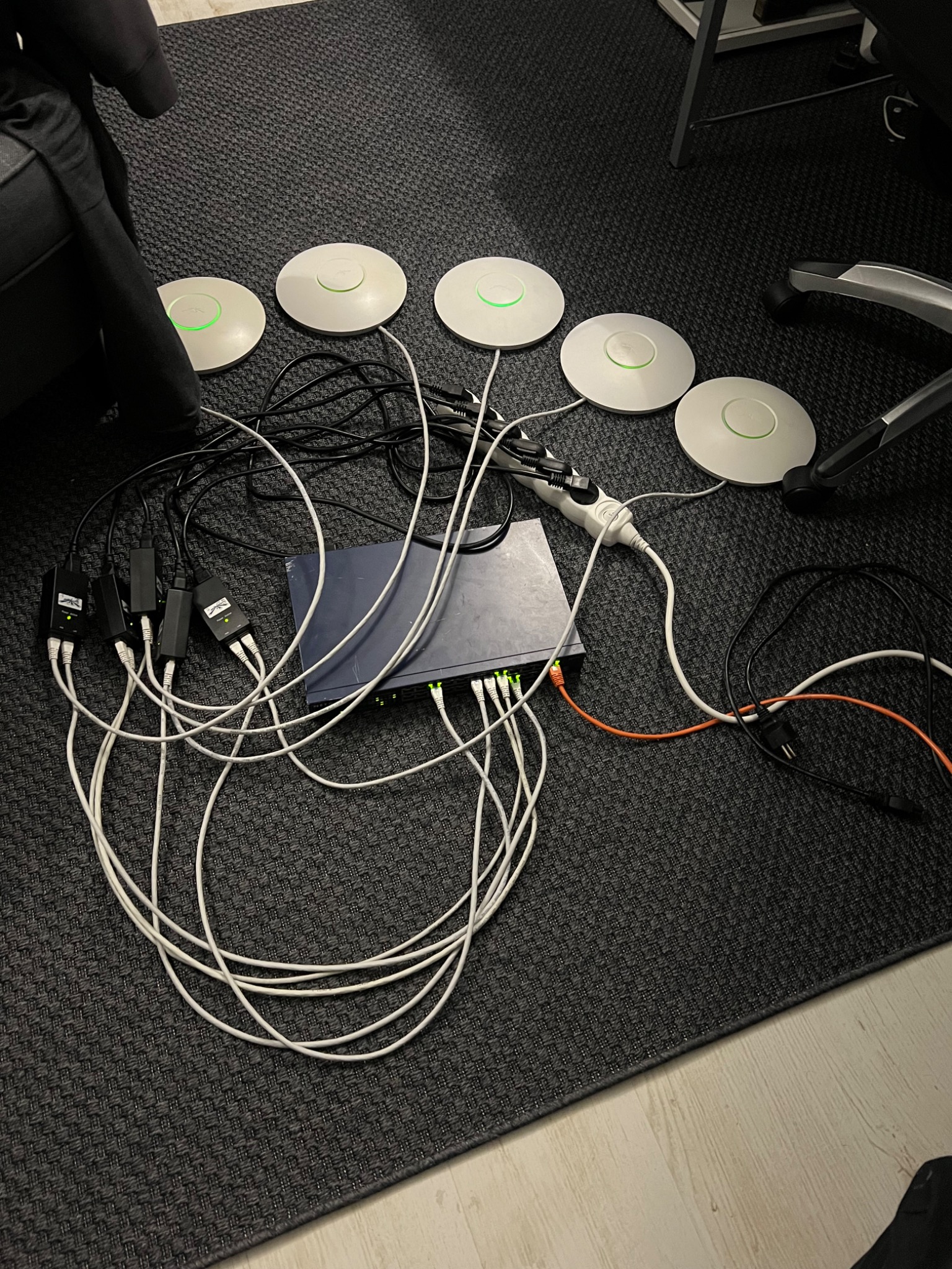 Rozłożone na ciemnoszarym dywanie: router, cztery białe punkty dostępowe z zielonymi diodami, zasilacze PoE i plątanina kabli sieciowych.