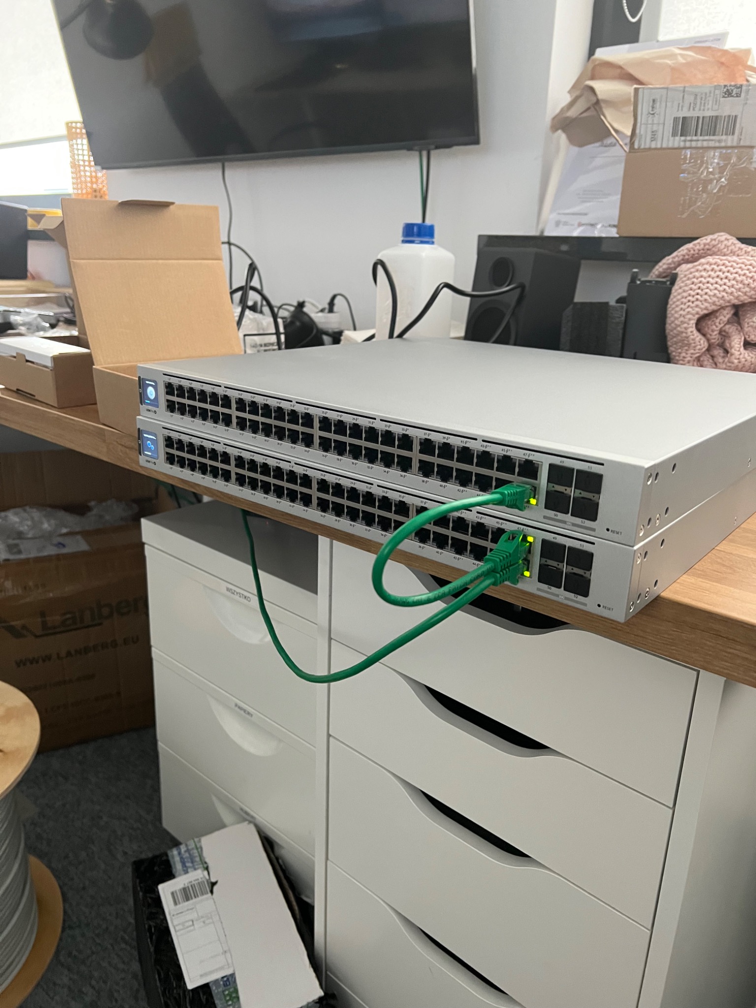 Dwa przełączniki sieciowe ustawione jeden na drugim, podłączone zielonymi kablami Ethernet, diody LED sygnalizują aktywność, tło stanowią biurko i biała komoda.