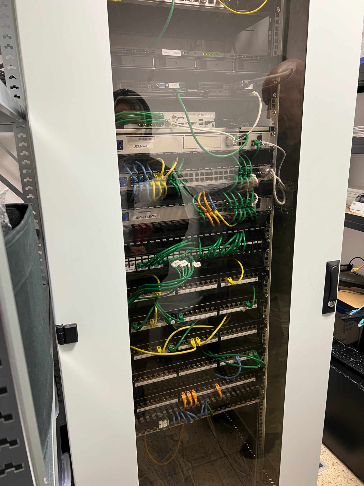 Serwerownia z szafą rack, widoczne switche, patch panele i okablowanie strukturalne w kolorach zielonym, żółtym i niebieskim. Przez szybę szafy widoczne odbicie osoby.