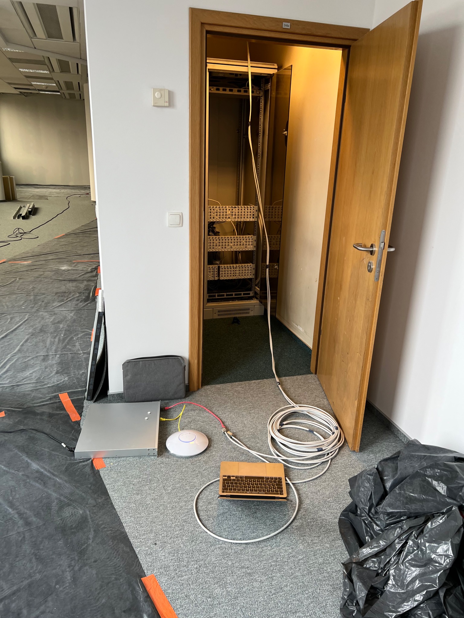 Instalacja sieci komputerowej w biurze: router, switch i laptop podłączone kablami przed otwartą szafą rack w trakcie pracy, podłoga zabezpieczona folią.