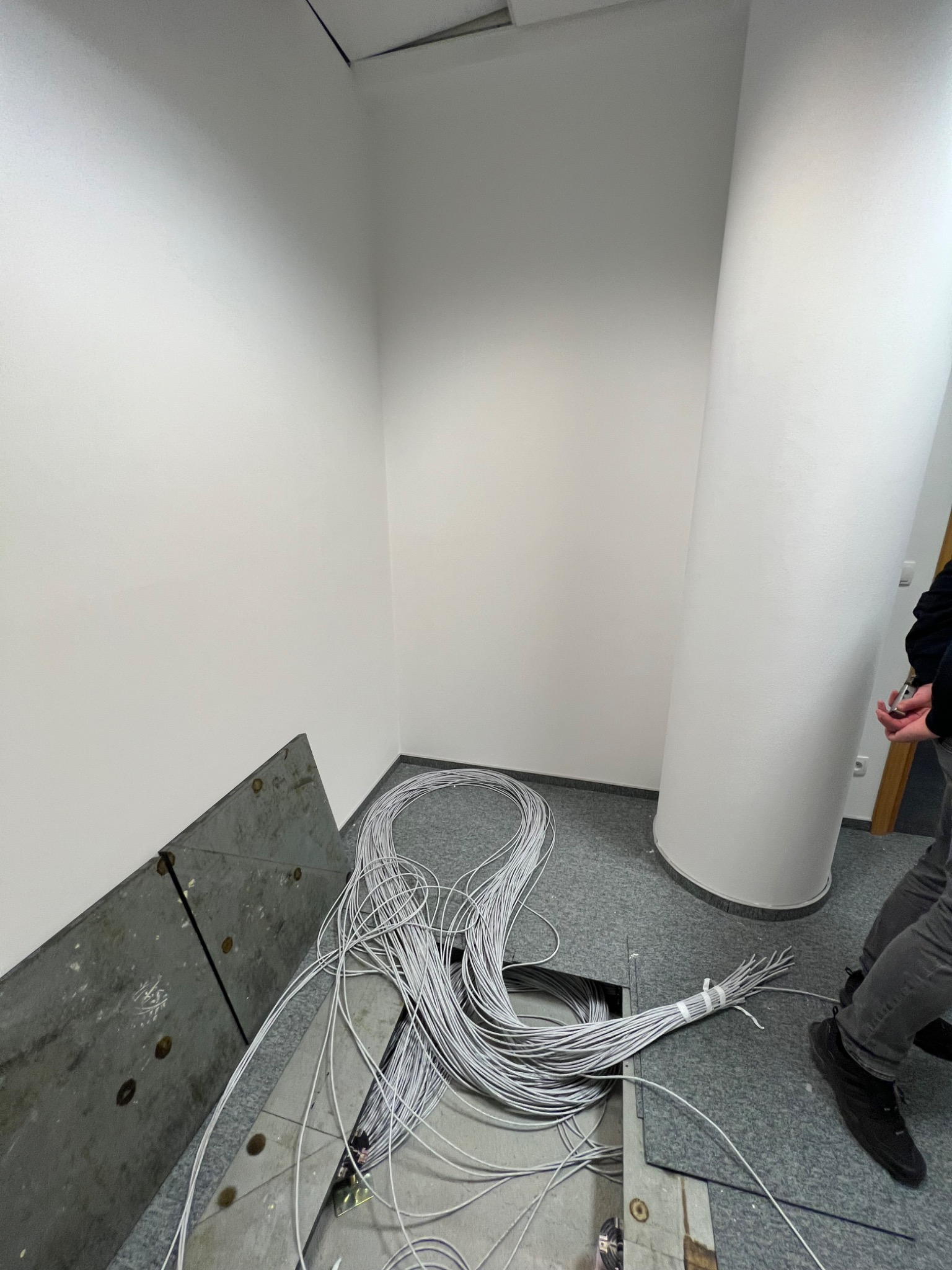 Rozłożona podłoga techniczna z widocznymi kablami, białe ściany i fragment kolumny, osoba w tle.