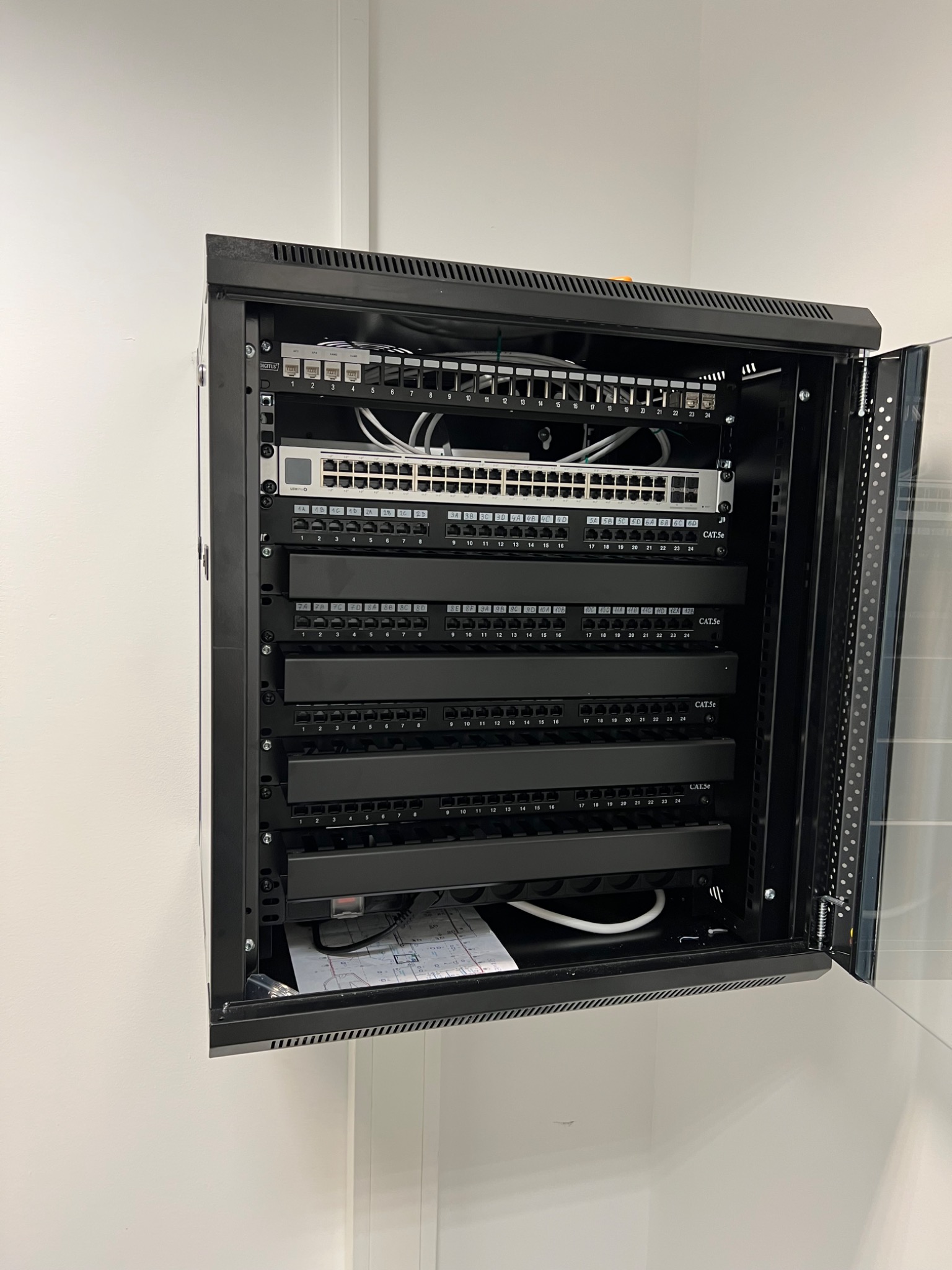 Otwarta szafa rack z panelem krosowniczym, switchem i okablowaniem CAT5e, widoczne porty oznaczone numerami, dokumentacja techniczna na dnie szafy.