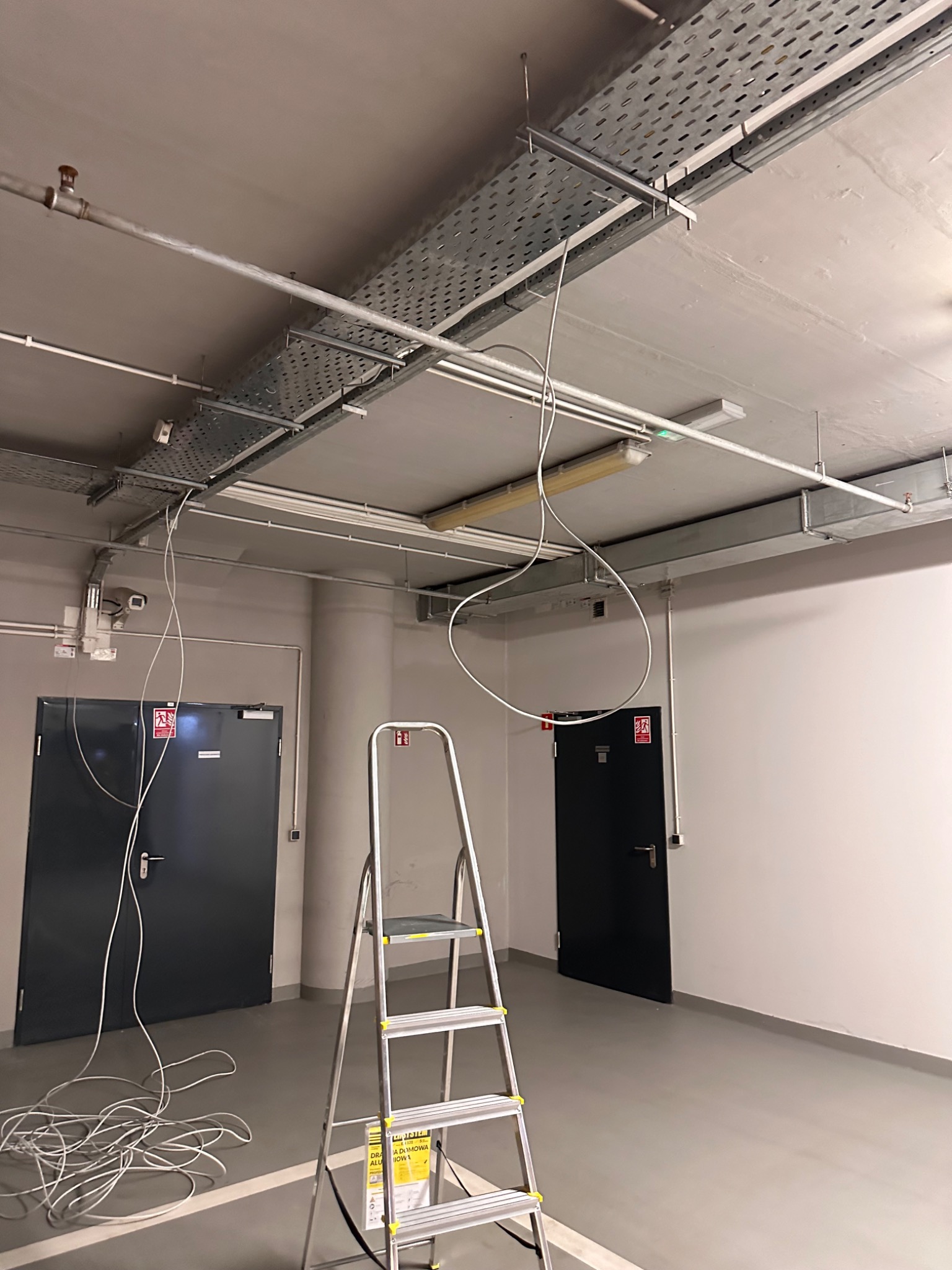 Wewnętrzne ujęcie z metalową drabinką stojącą na szarym podłożu, kable zwisające z perforowanych korytek kablowych pod sufitem, dwie ciemne drzwi po bokach i białe ściany, sugerujące prace...