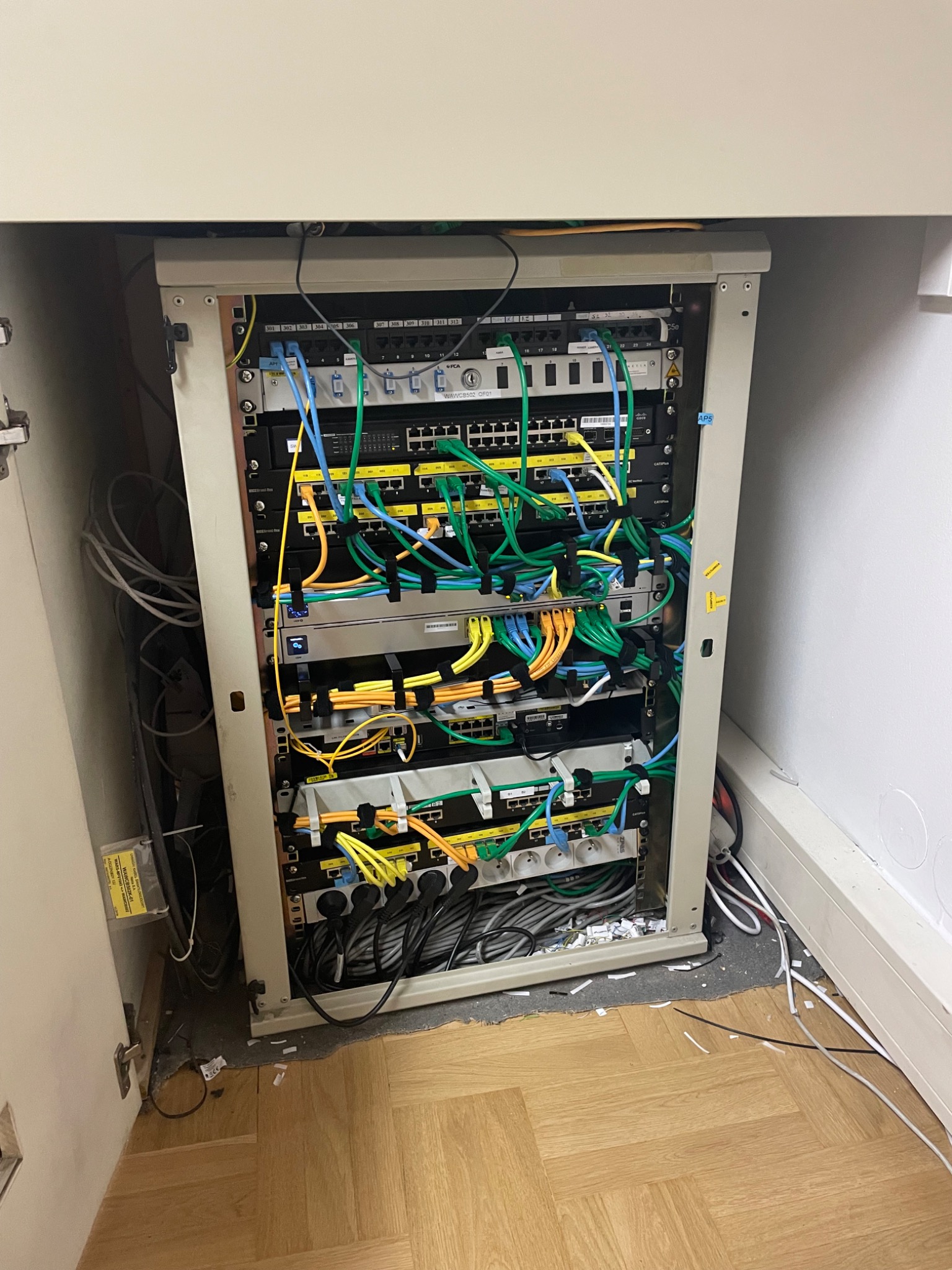 Szafa rack z zainstalowanymi przełącznikami sieciowymi, patch panelem i listwami zasilającymi, okablowanie strukturalne w kolorach niebieskim, zielonym i żółtym, widoczne pozostałości po instalacji...