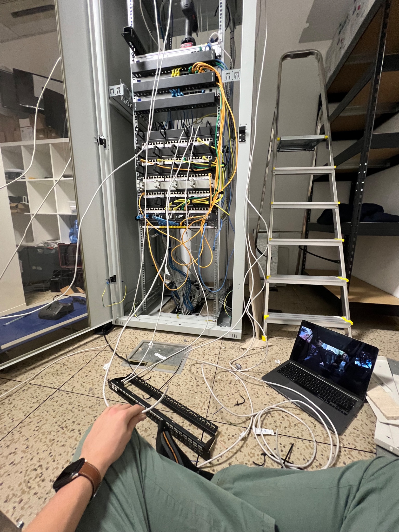 Otwarta szafa serwerowa z widocznymi patch panelami i switchami, niepodłączone kable, drabina aluminiowa, laptop z włączonym filmem i ręka z zegarkiem na pierwszym planie.