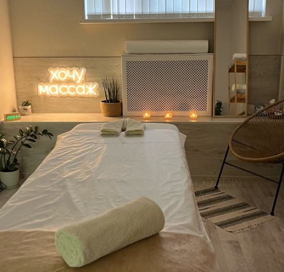 Jasne wnętrze gabinetu z leżanką do zabiegów, podświetlonym napisem neonowym 'Chcę masaż', świecami i roślinami. Przytulna atmosfera.