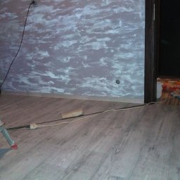 Daniel Dychus - Ściana pomalowana w nieregularne wzory w odcieniach szarości i bieli, widoczne gniazdka elektryczne, podłoga z paneli imitujących drewno, fragment drabiny malarskiej i przewód zasilający na podłodze.