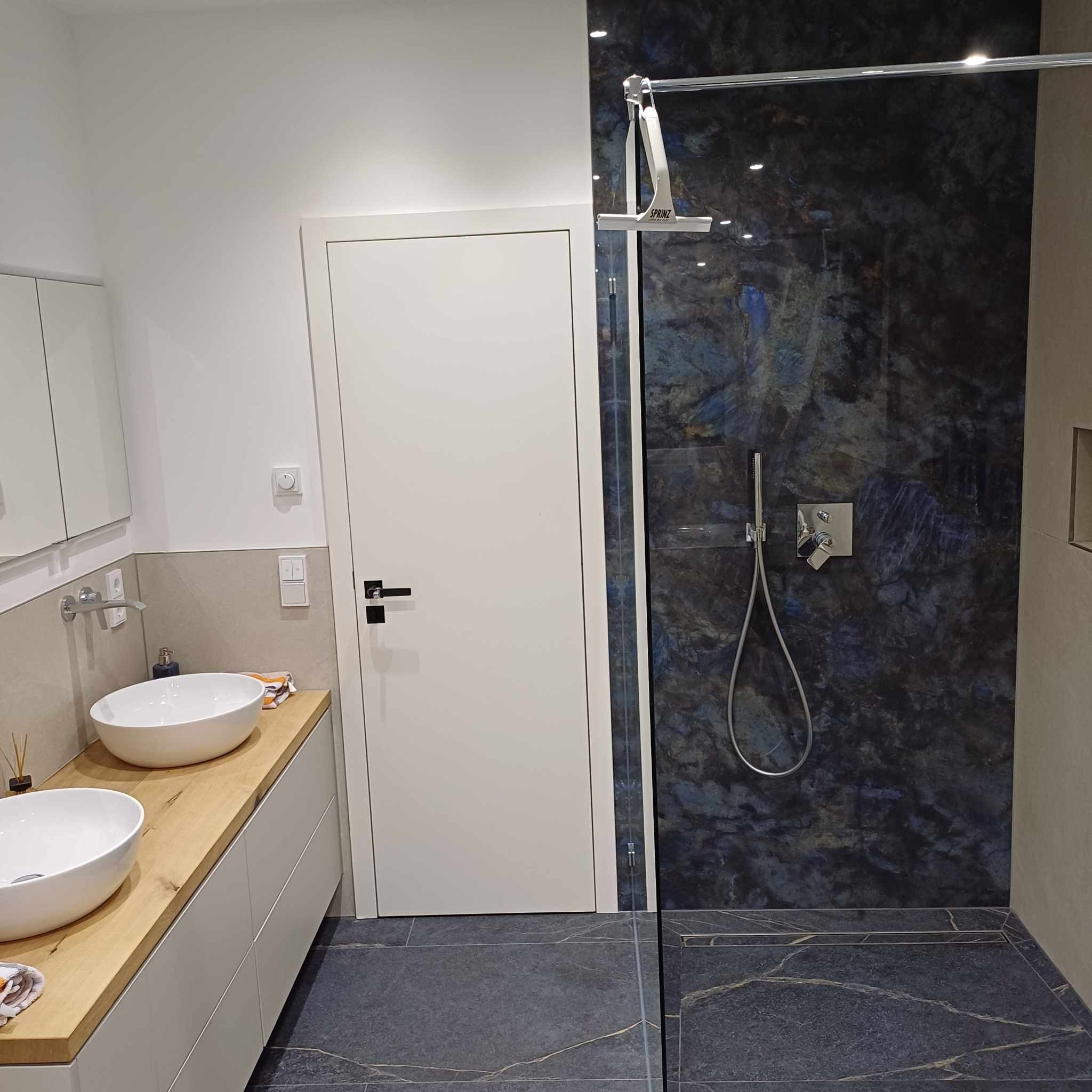 Nowoczesna łazienka z dwoma białymi umywalkami na drewnianym blacie, kabina prysznicowa z ciemną, marmurkową ścianą i chromowaną armaturą, białe drzwi i szara podłoga z odpływem liniowym.