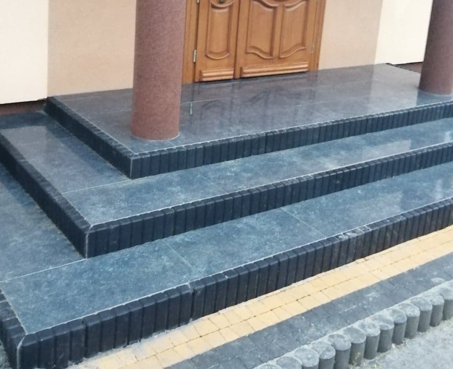 Zewnętrzne schody wejściowe wyłożone szarymi płytkami z ciemnymi obrzeżami, prowadzące do drewnianych drzwi wejściowych, wsparte na brązowych, okrągłych kolumnach.