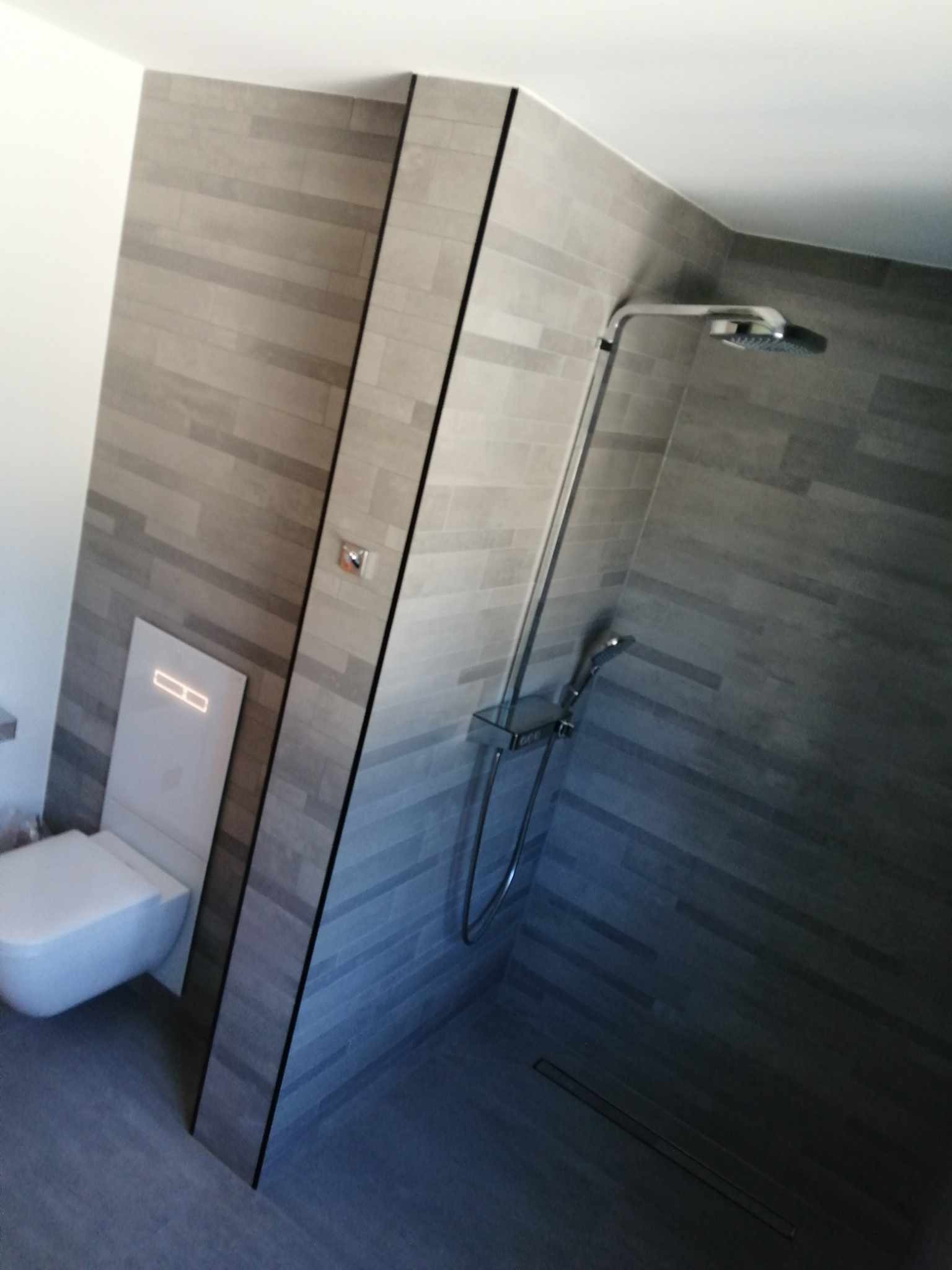 Nowoczesna łazienka z szarymi płytkami imitującymi drewno, minimalistyczna toaleta podwieszana z podświetleniem LED oraz chromowana bateria prysznicowa z deszczownicą.