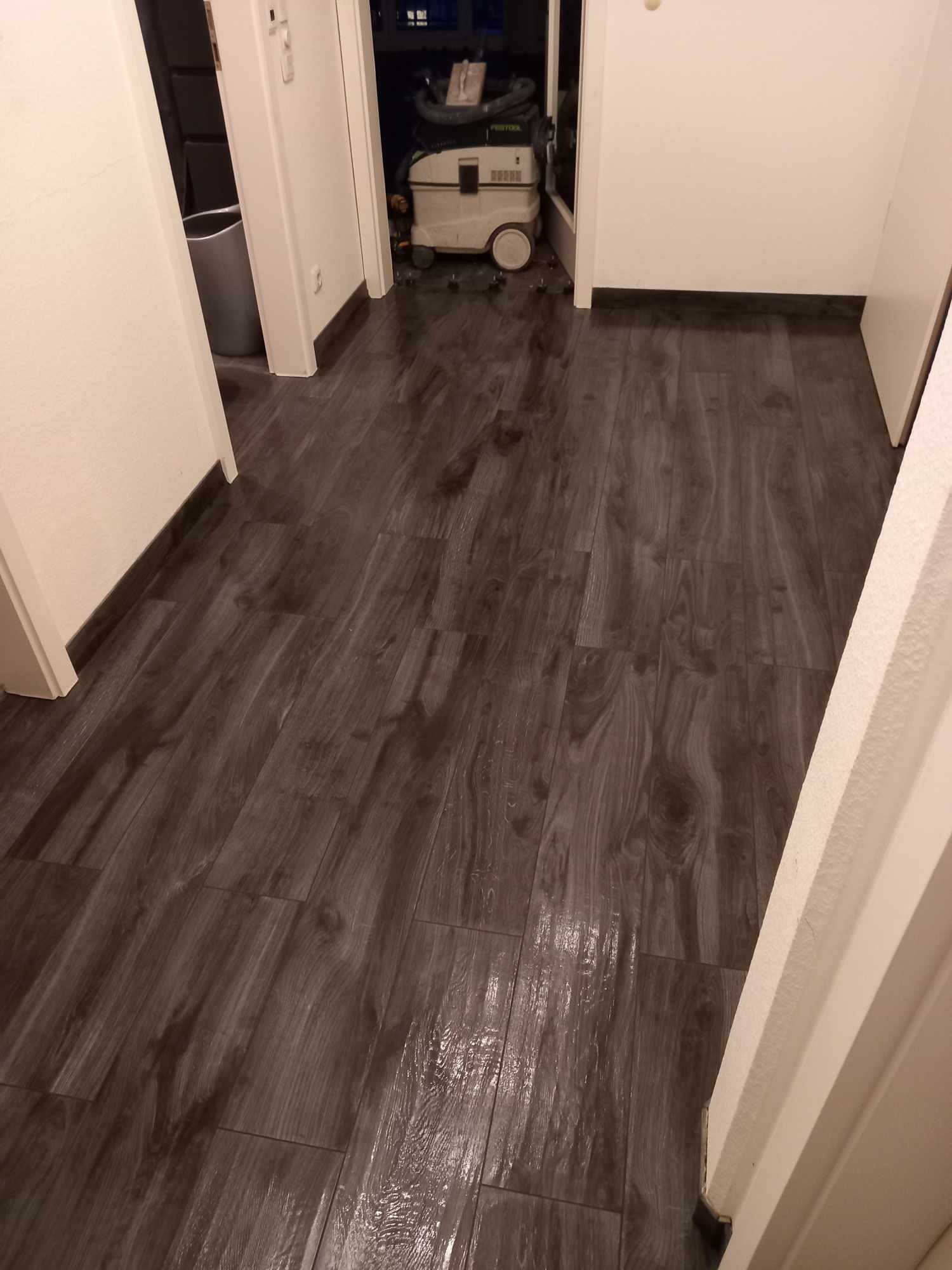 Ułożone panele podłogowe w odcieniach brązu, imitujące drewno, z widocznym odkurzaczem przemysłowym w tle i przejściem do innego pomieszczenia.