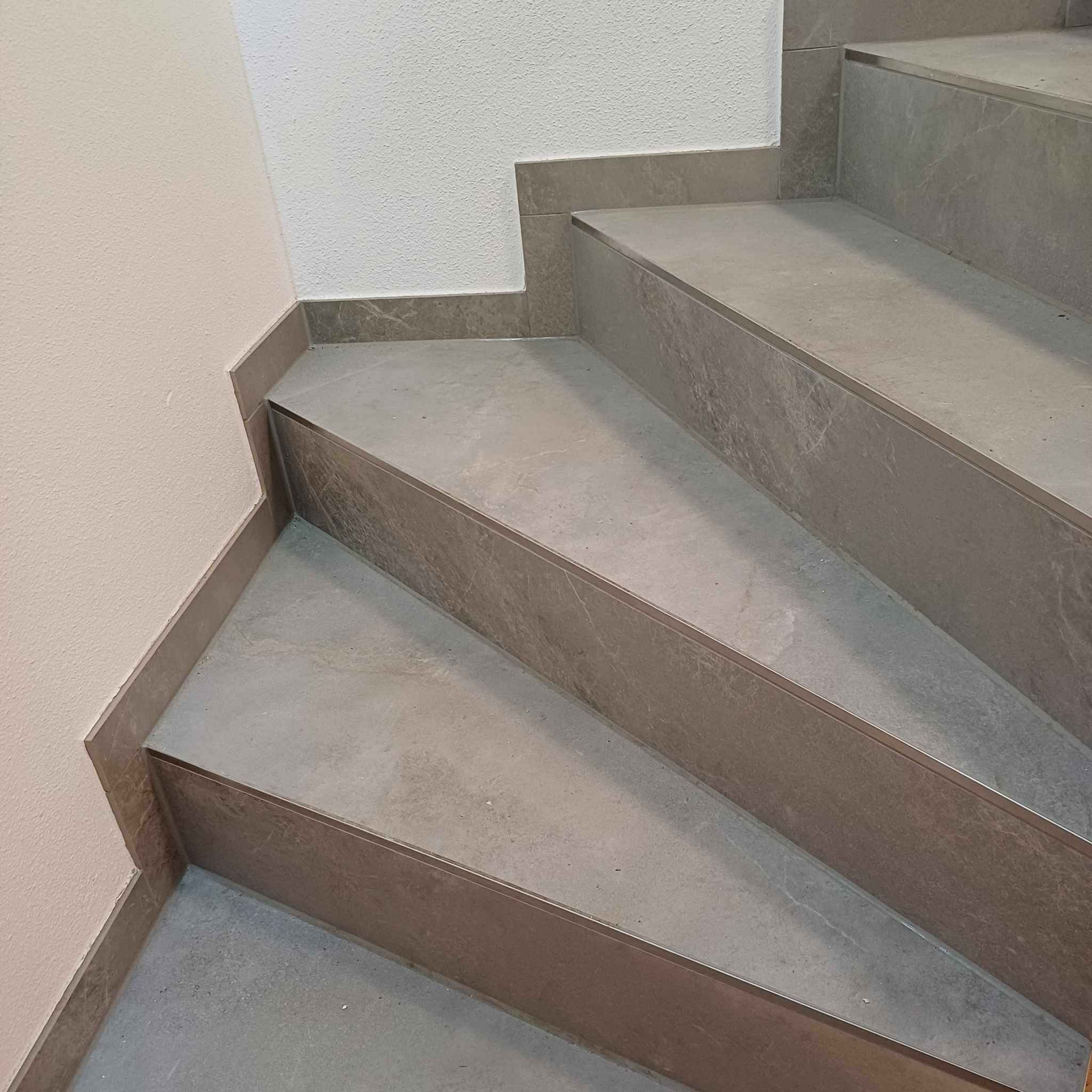 Wykończone schody wewnętrzne z szarych płytek imitujących marmur, z metalowymi listwami na krawędziach stopni, przy ścianie pomalowanej na dwa odcienie bieli i beżu.