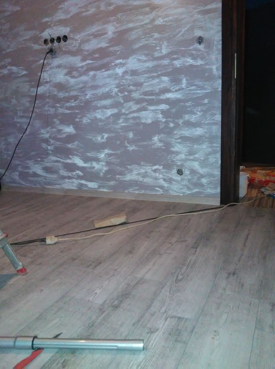 Ściana pomalowana w nieregularne wzory w odcieniach szarości i bieli, widoczne gniazdka elektryczne, podłoga z paneli imitujących drewno, fragment drabiny malarskiej i przewód zasilający na podłodze.