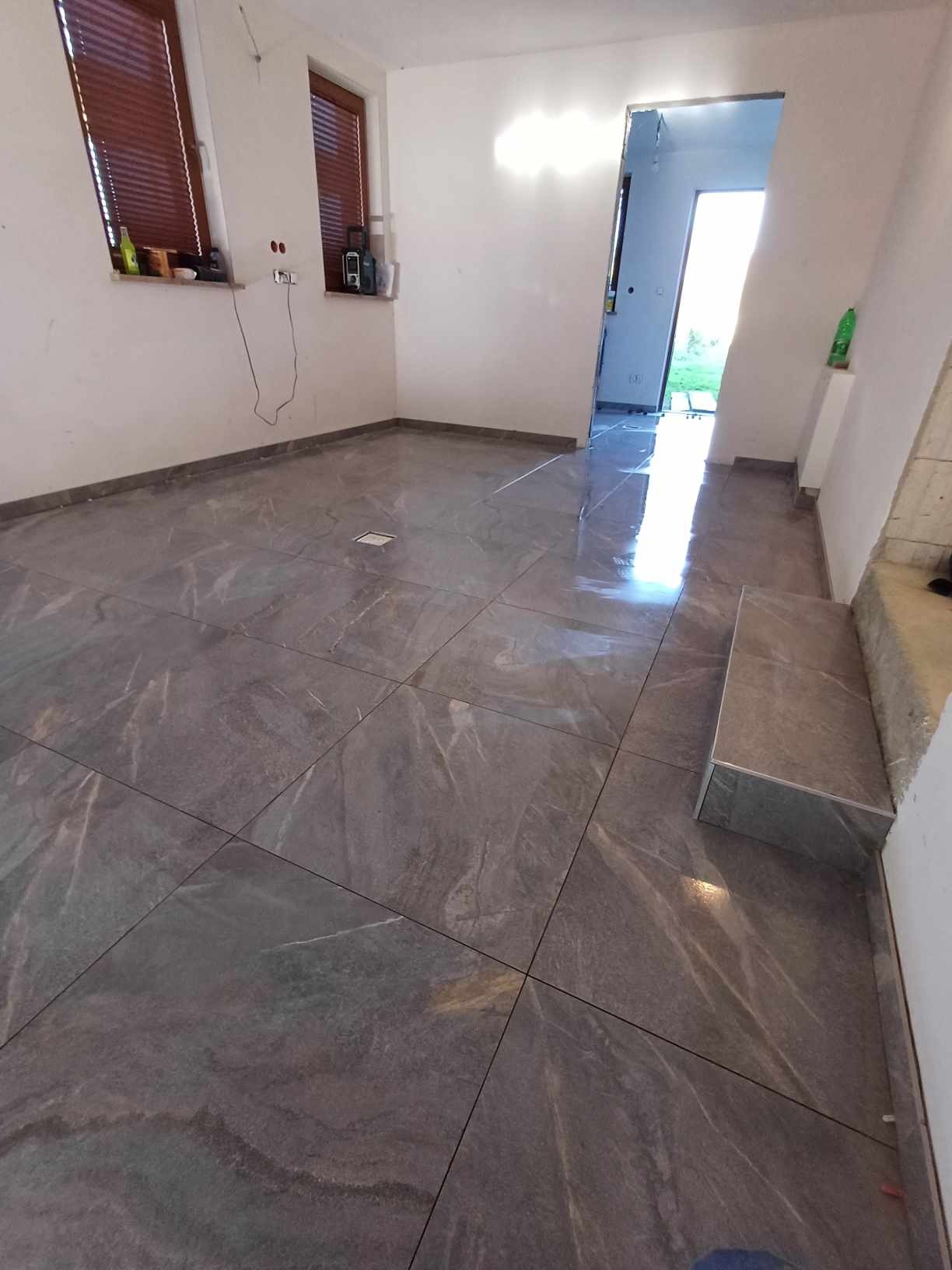 Wykończona podłoga z płytek imitujących kamień, z widocznym odpływem liniowym i progiem, w jasnym, odnowionym pomieszczeniu z dwoma oknami.