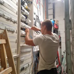 Elektryk w trakcie pracy przy rozbudowanej skrzynce rozdzielczej z bezpiecznikami i okablowaniem, używa śrubokręta, drabina drewniana z lewej strony, widoczne przewody i elementy instalacji.