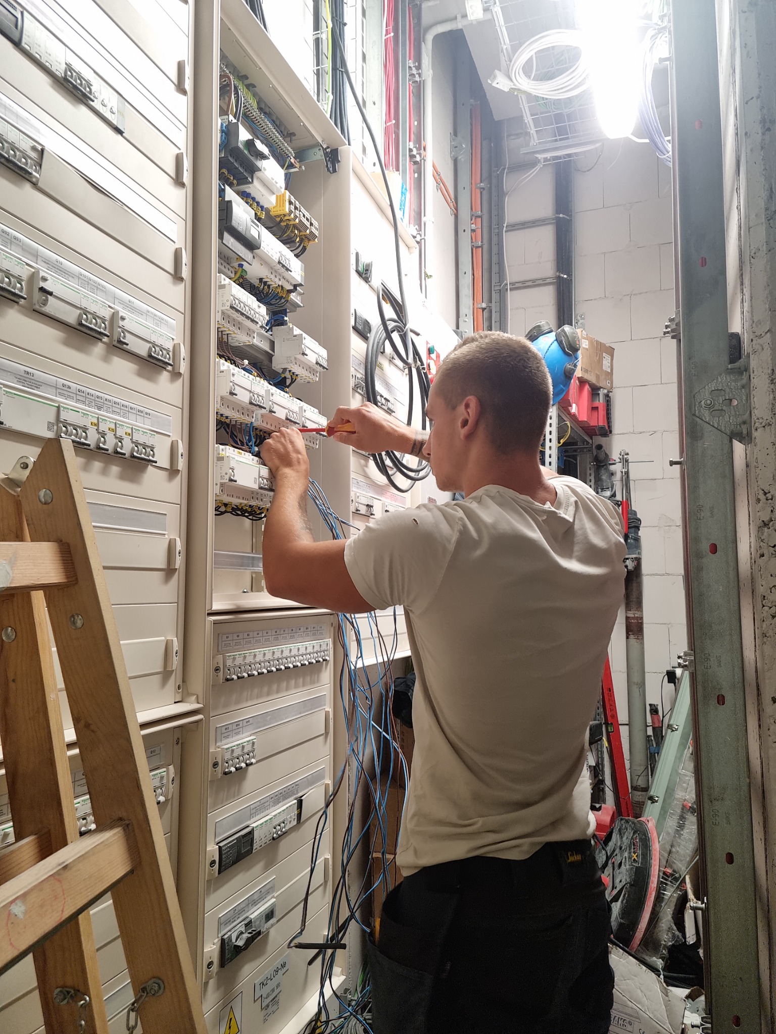 Elektryk w trakcie pracy przy rozbudowanej skrzynce rozdzielczej z bezpiecznikami i okablowaniem, używa śrubokręta, drabina drewniana z lewej strony, widoczne przewody i elementy instalacji.
