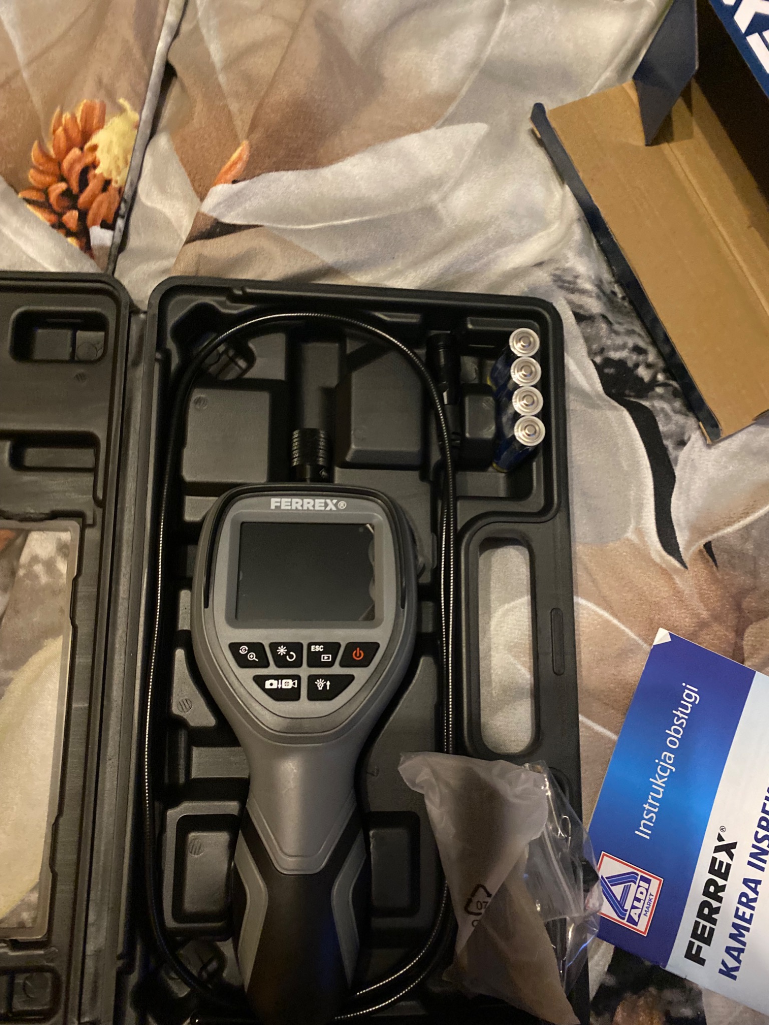 Kamera inspekcyjna Ferrex w czarnym futerale z czterema bateriami AA i instrukcją obsługi na wzorzystej pościeli.