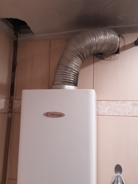 Biały piec gazowy marki Euroterm z podłączonym, giętkim przewodem odprowadzającym spaliny do komina, widoczne płytki ceramiczne na ścianie.