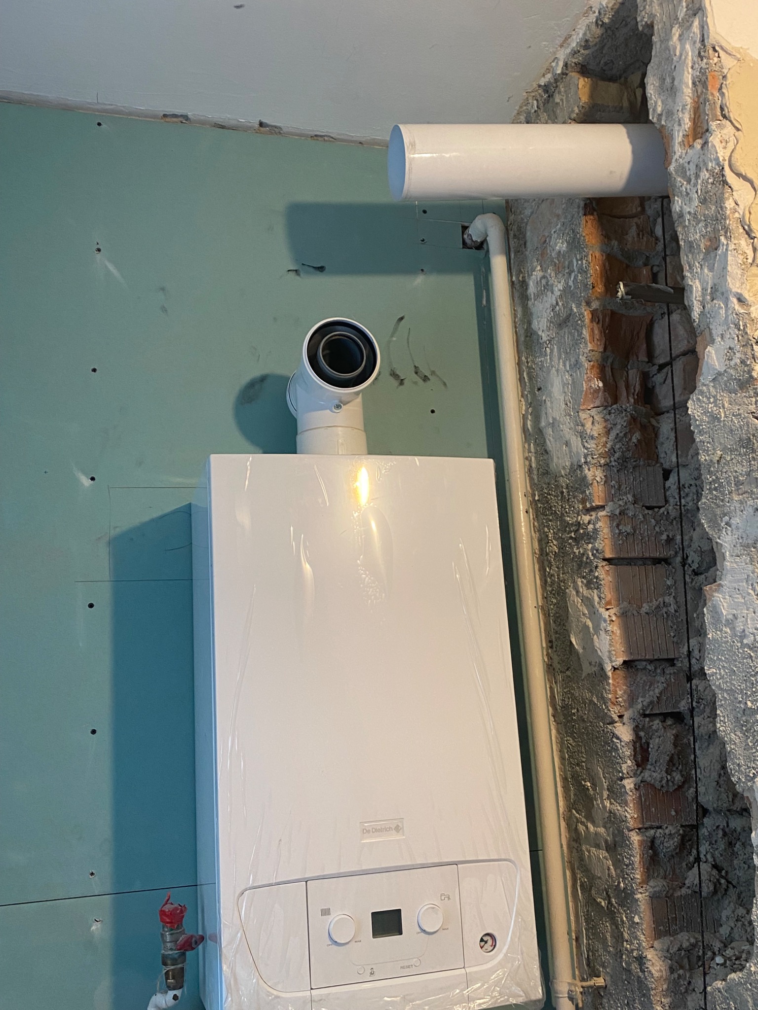 Nowy, biały piec gazowy De Dietrich w folii ochronnej, z podłączonymi rurami wentylacyjnymi, zamontowany na ścianie z płyt gipsowo-kartonowych i ceglanym murem w trakcie wykończenia.