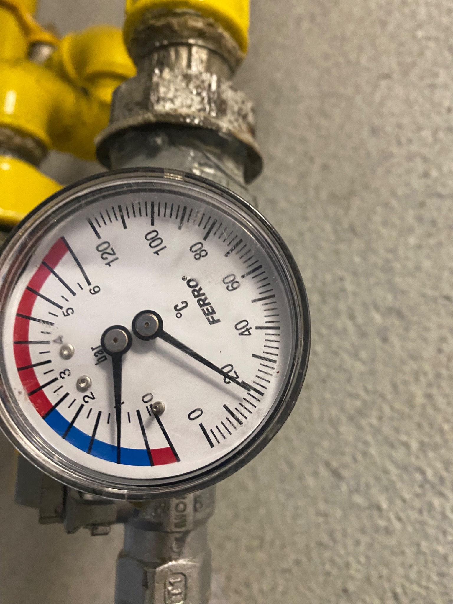 Manometr Ferro z podziałką temperatury i ciśnienia, zamontowany na metalowej instalacji z żółtymi elementami, wskazujący temperaturę pokojową.