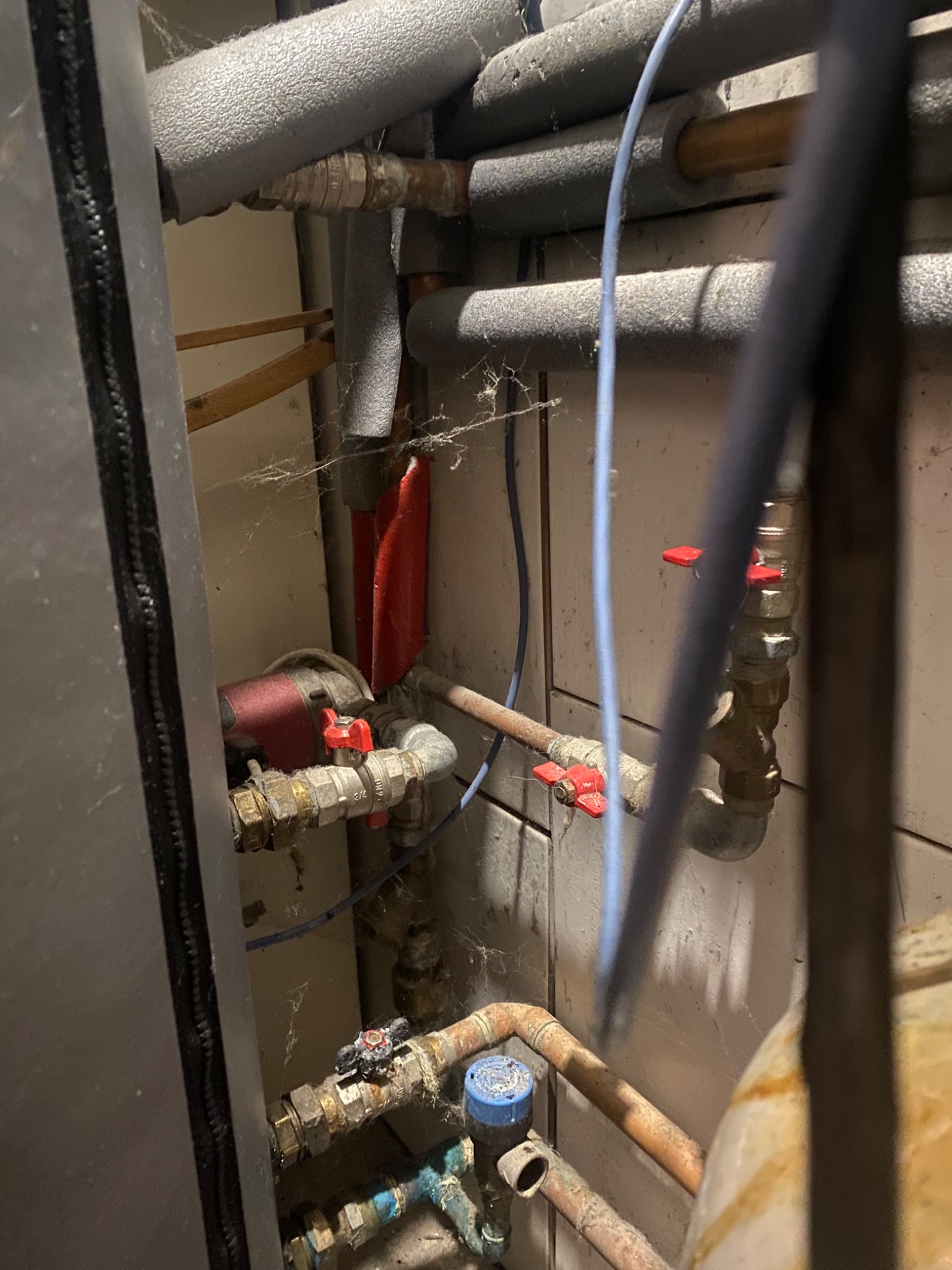 Fragment instalacji grzewczej z licznymi rurami, zaworami z czerwonymi pokrętłami i elementami izolacji, pokrytymi pajęczynami, na tle białych płytek.