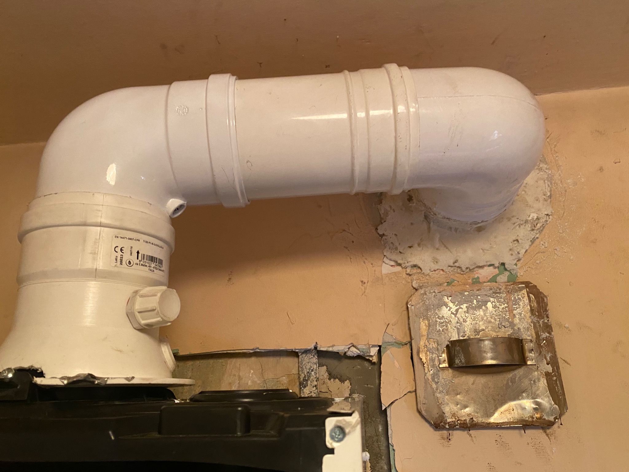 Biały, plastikowy przewód spalinowy pieca gazowego, zamontowany do ściany z widocznymi śladami napraw i starej farby. Widoczne elementy pieca i fragment metalowej osłony na ścianie.