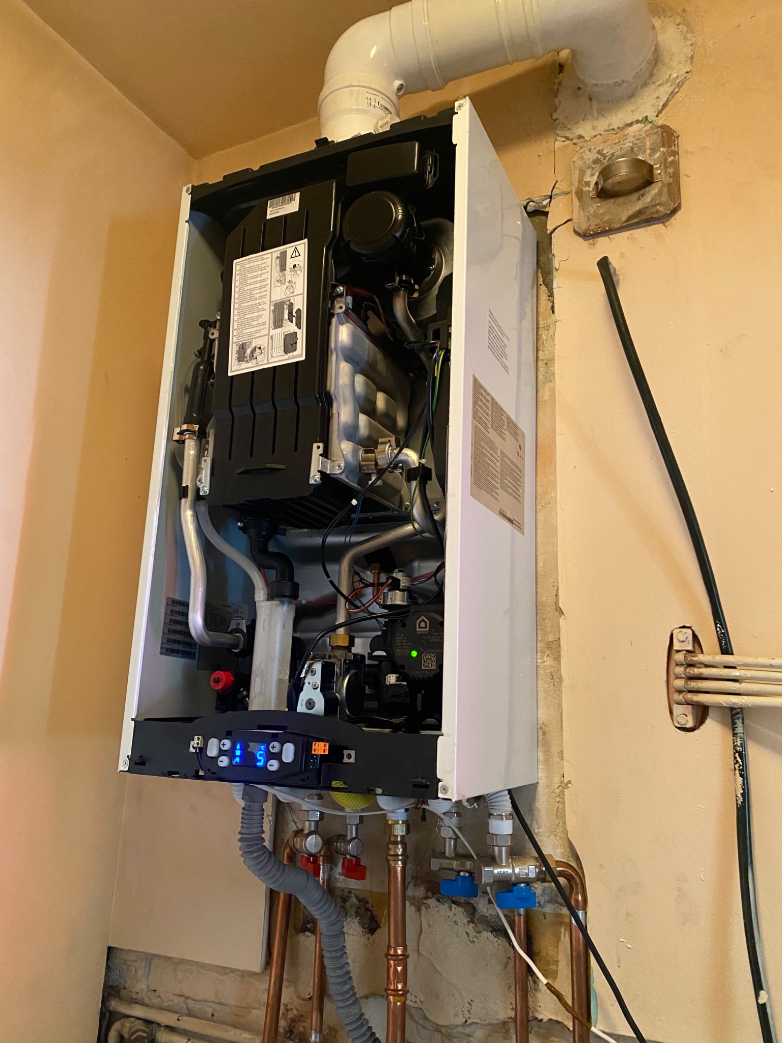 Nowoczesny piec gazowy zamontowany na ścianie z widocznymi rurami miedzianymi, zaworami oraz wyświetlaczem z niebieskim podświetleniem.