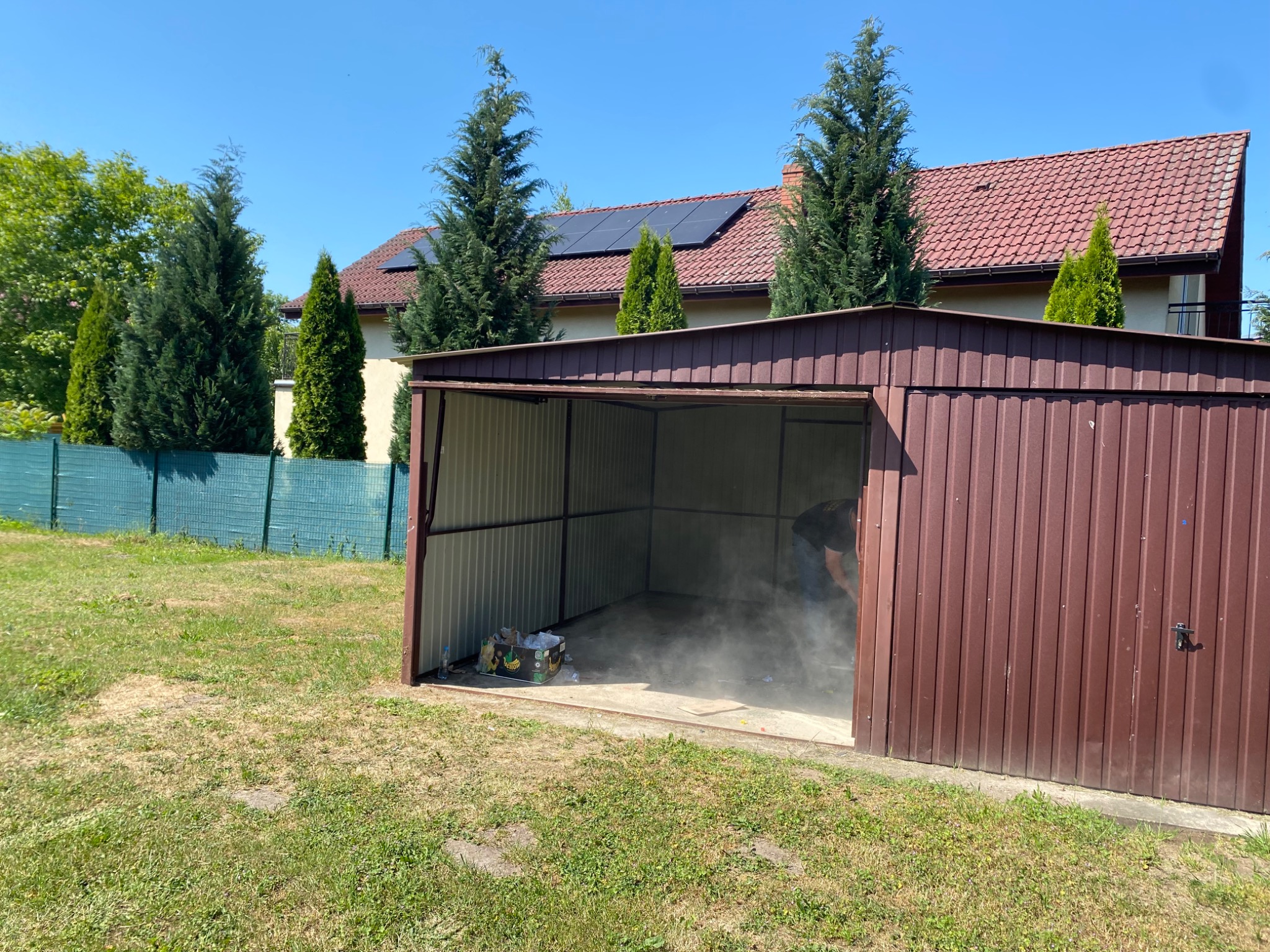 Metalowy garaż z otwartymi drzwiami, wewnątrz widoczny kurz i osoba w trakcie sprzątania, na zewnątrz trawnik i dom z panelami słonecznymi na dachu.