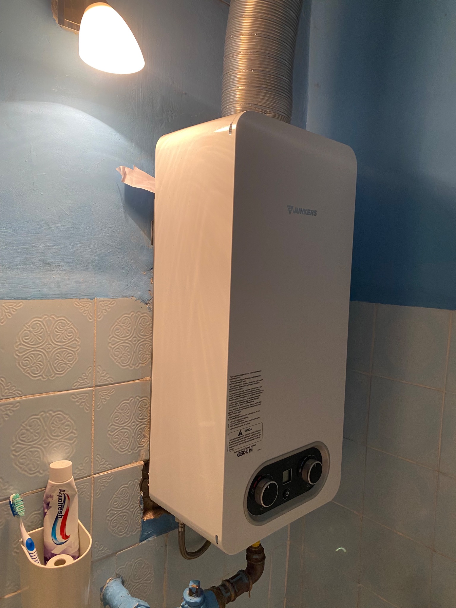 Biały piec gazowy Junkers zamontowany na niebieskiej ścianie z kafelkami, widoczny przewód odprowadzający spaliny, pod spodem rury instalacji oraz pojemnik ze szczoteczką i pastą do zębów.