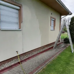 Fragment elewacji domu z widocznym śladem po umyciu, drabina oparta o ścianę, żółty wąż ogrodowy podłączony do kranu, kostka brukowa na ścieżce, trawnik i klimatyzator na ścianie.