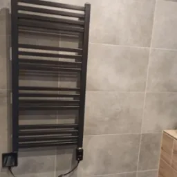Czarny, elektryczny grzejnik łazienkowy drabinkowy zamontowany na szarej ścianie z płytek ceramicznych.