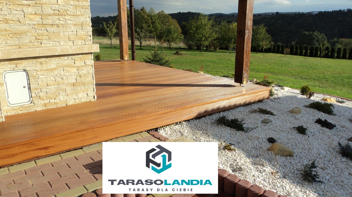 Wykończony taras z drewna egzotycznego, z widocznym logo firmy Tarasolandia, otoczony białym żwirem i zielenią, na tle domu z kamienia.