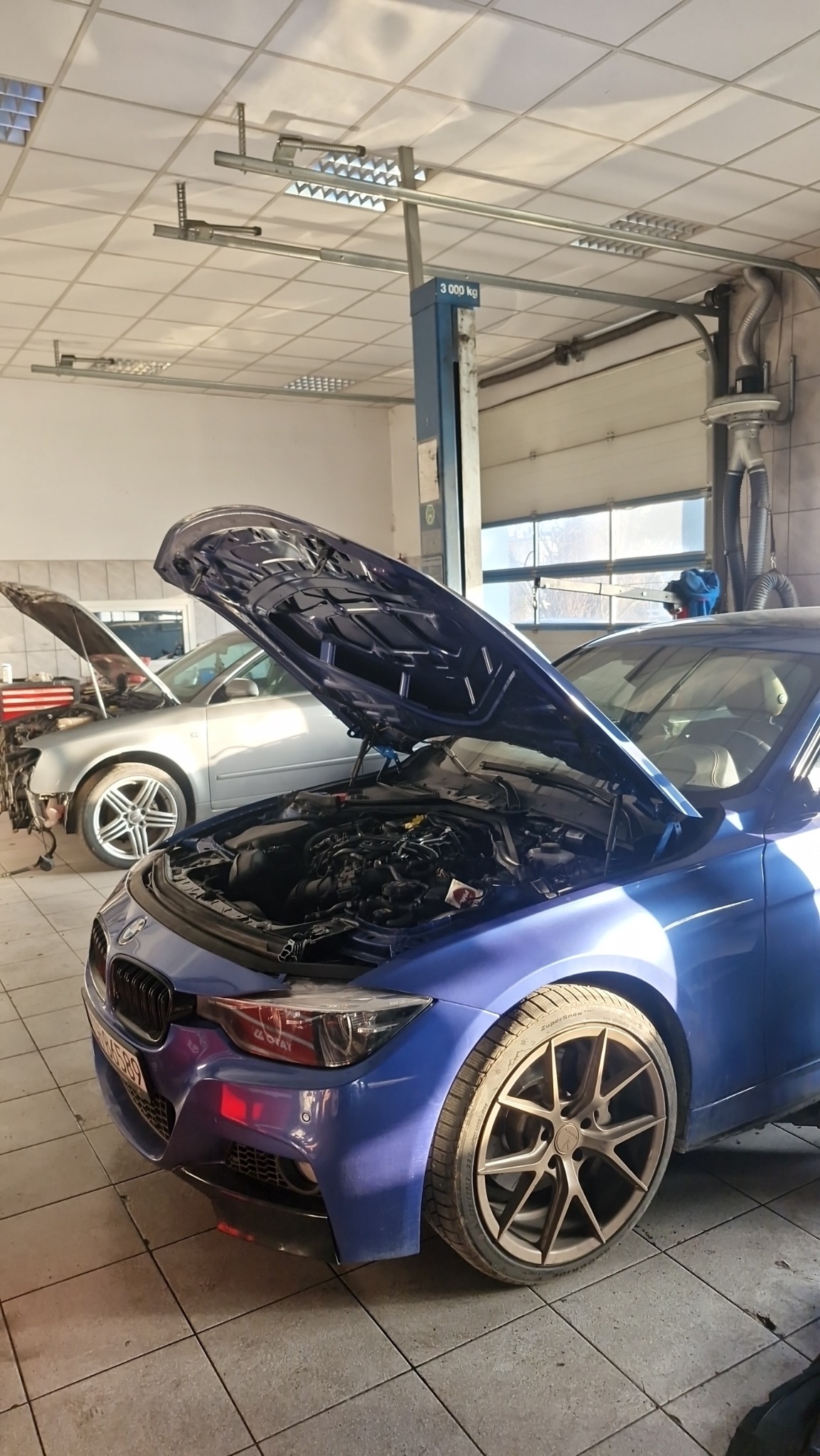 Niebieskie BMW z otwartą maską w warsztacie; widoczny silnik i inne auto w tle z podniesioną maską, oświetlenie warsztatowe.