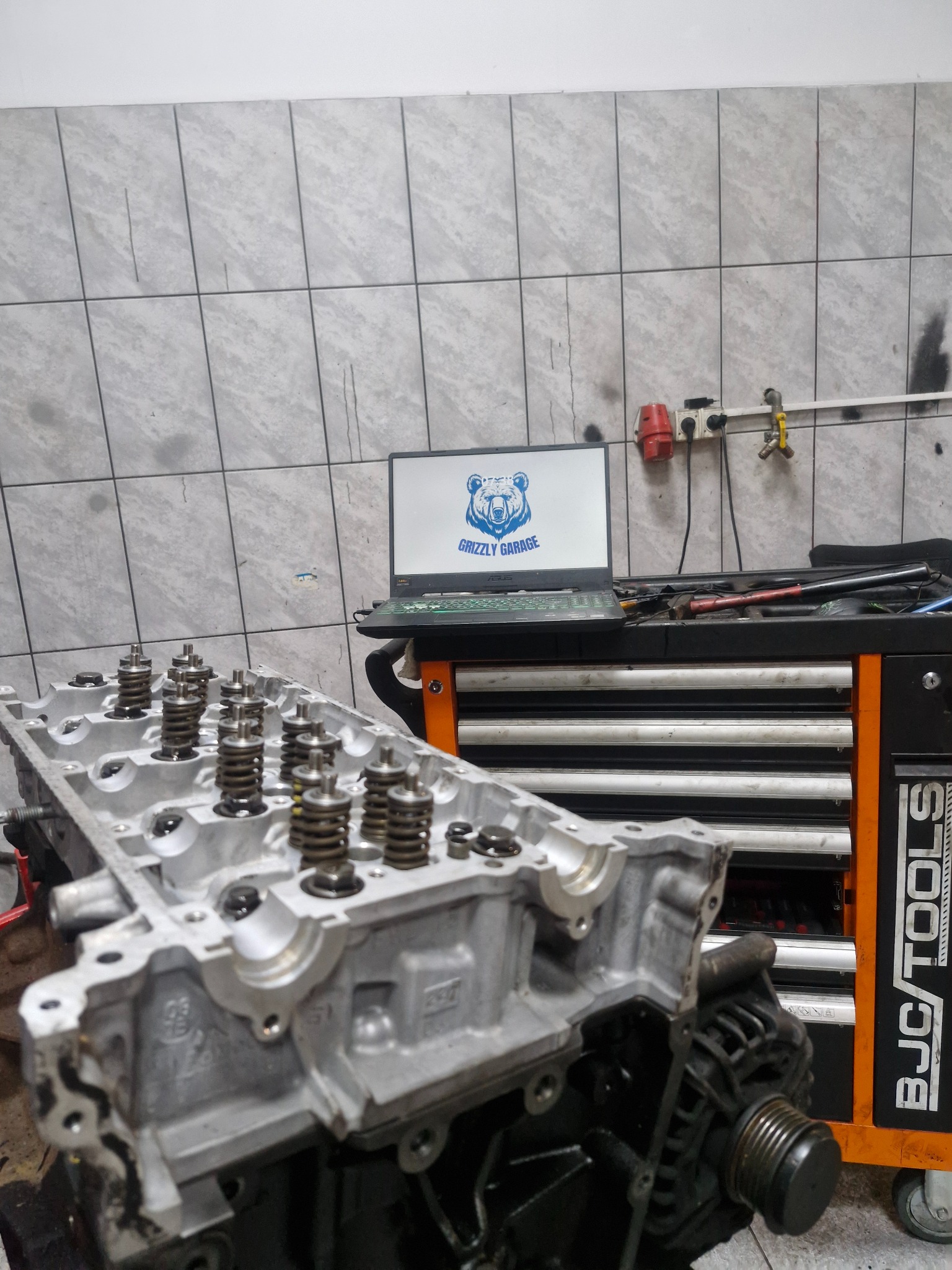 Głowica silnika z widocznymi sprężynami zaworowymi na tle warsztatu z laptopem wyświetlającym logo 'Grizzly Garage' i skrzynką narzędziową BJC/Tools.