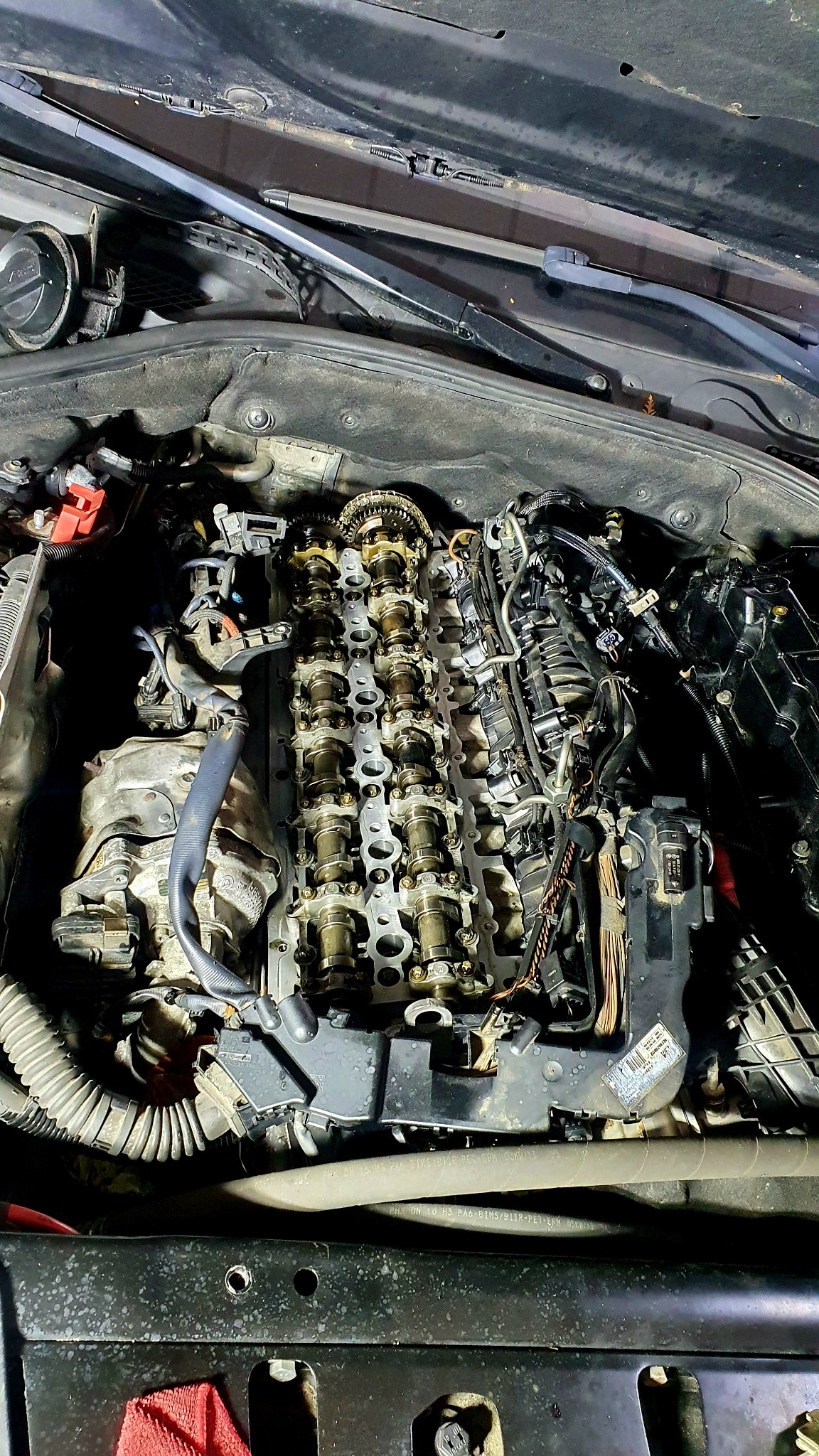 Otwarty silnik samochodowy z widocznymi wałkami rozrządu i łańcuchem, w trakcie naprawy, z elementami osprzętu i przewodami. Widoczna czerwona ściereczka w dolnej części zdjęcia.
